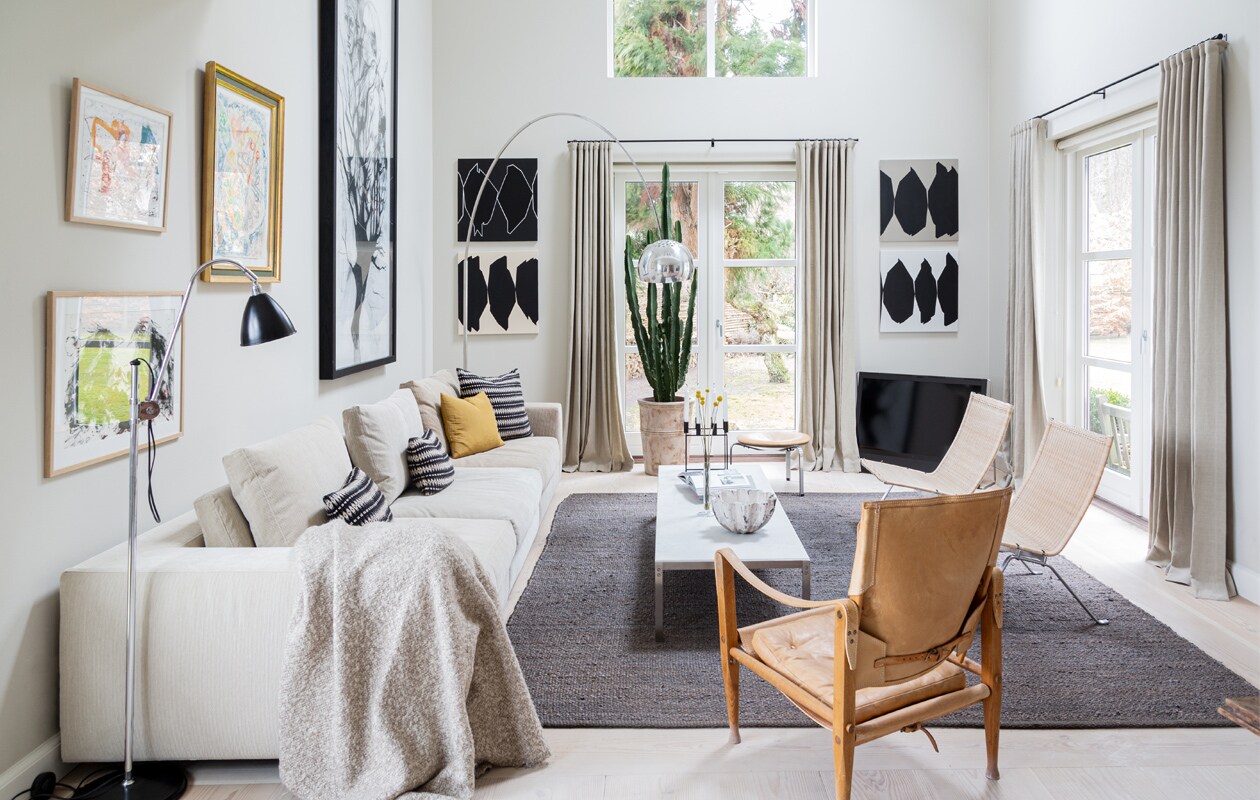 Muurverf Shaded White van Farrow &amp; Ball. Bank Eilersen. Kussens Tapet-Cafe. Koffietafel PK62 en rotan stoelen PK22 van Poul Kjærholm voor Fritz Hansen. Leren stoel Kaare Klint voor Carl Hansen &amp; Son. Vloerlamp Arco van Flos. Schaal gemaakt door een vriendin. Kaarsenstandaard Mogens Lassen. Schilderijen van links naar rechts Carsten Dahl (twee maal), Eigill Jacobsen, Cathrine Raben Davidsen. Grafische kunstwerken Ulla Pedersen. Vloerkleed Tine K Home.