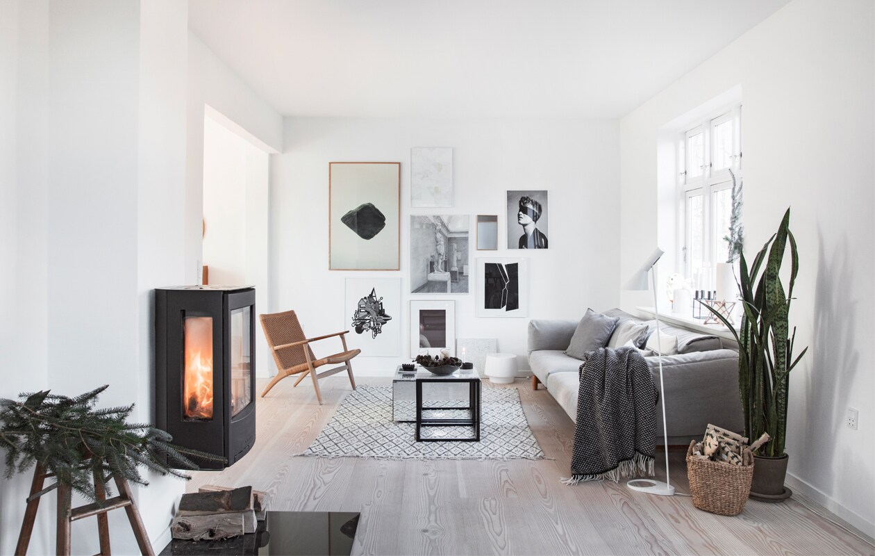 Op de vintage trapkruk dennentakken uit de tuin. Houtkachel Wiking. Bank Muuto. Tafels Kristina Dam Studio. Hans Wegner fauteuil Fritz Hansen. Kleed Nordal. Vloerlamp AJ Louis Poulsen. Plaid Hübsch. Mand Intratuin. Plant Dracaena trifasciata (Vrouwentong) in pot. Kunstwerken mix van zwart-witfoto’s en deels zelfgemaakte illustraties.