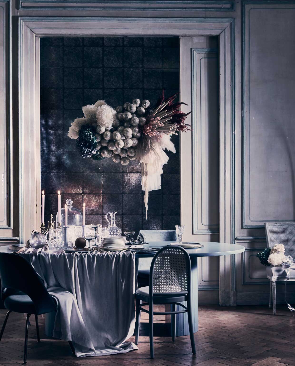 Wolk van bloemen en glitterballen, op maat gemaakt door Edenique Floral Design, prijs op aanvraag (edenique.nl). Eettafel vanaf € 800,- (com-anders.nl). Grijze fluwelen eetkamerstoel € 1210,-, grijze stoel van Thonet met fluwelen zitting € 895,-, grijze fluwelen stoel rechtsachter, vanaf € 1200,- (edha-interieur.nl). Wit met grijs linnen beklede Franse antieke webbing stoel, prijs op aanvraag, grote glazen stolp , € 195,- (pietjonker.nl). Glazen karaffen, vanaf € 60,-, tweearmige kandelaar, prijs op aanvraag (prinsheerlijkantiek.nl). Grijze glazen champagnecoupes van &amp;klevering, set van 2 € 27,50, goudkleurig bestek 16-delig € 149,-,  (vtwonen.nl). Wijnglas in kristallook , € 7,99, witte borden met gouden rand, Ø 21 cm € 7,99, keramieken kandelaar € 17,99 (hm.com). Glazen kerstbal € 9,75 (iblaursen.nl). Zwart-witte borden met gezichten € 115,- , goudkleurige onderborden vanaf € 30,-,  (les-ottomans.com). Tafelkleed van fluwelen stof Velveto FRC2400/07 van Designers Guild, 140 cm breed, per meter € 141,34 (wva.nl). Behang Kings Mirror 98/13055 van Cole &amp; Son, per rol 50 cm x 10 m € 128,- (deruamsterdam.nl). Okerkleurige kerstbal € 4,50 (nordal.eu). Overig privébezit., , ,