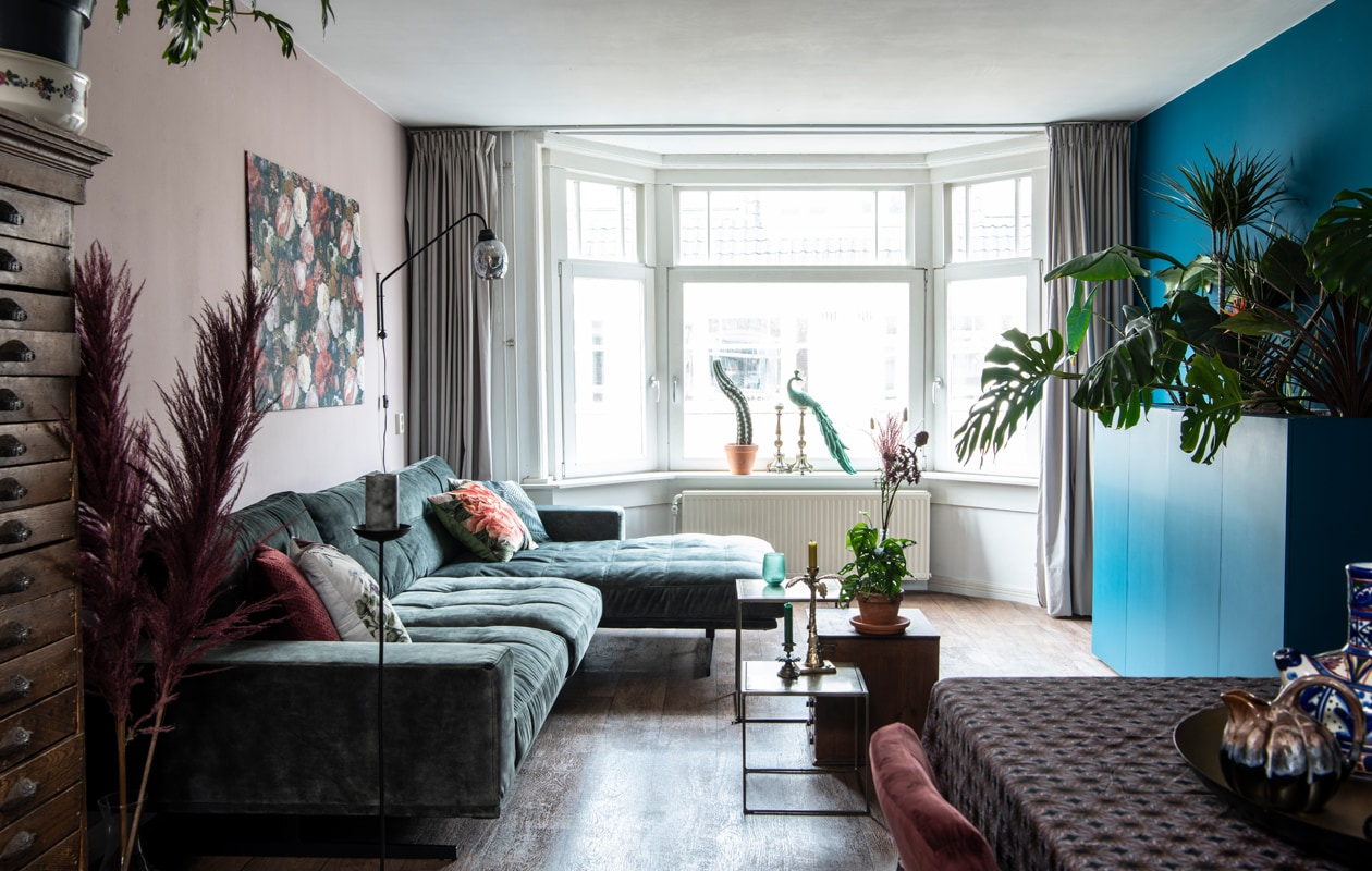 Bank, bijzettafels, cactus, plant op ladekast, kaarsenstandaards op tafel, schaal en veren Blooming Interior. Kussens Vanilla Fly, lamp BePureHome. Pauw By-Boo. Ladekast gemaakt door de opa van een vriend. Herkomst gordijnen onbekend. Kaarsenstandaards voor het raam IJ-Hallen. Het zeil lag er al. Muurverf Light Peachblossom 3 van Little Greene. Kunstwerk zelfgemaakt. Gordijnen Hema.