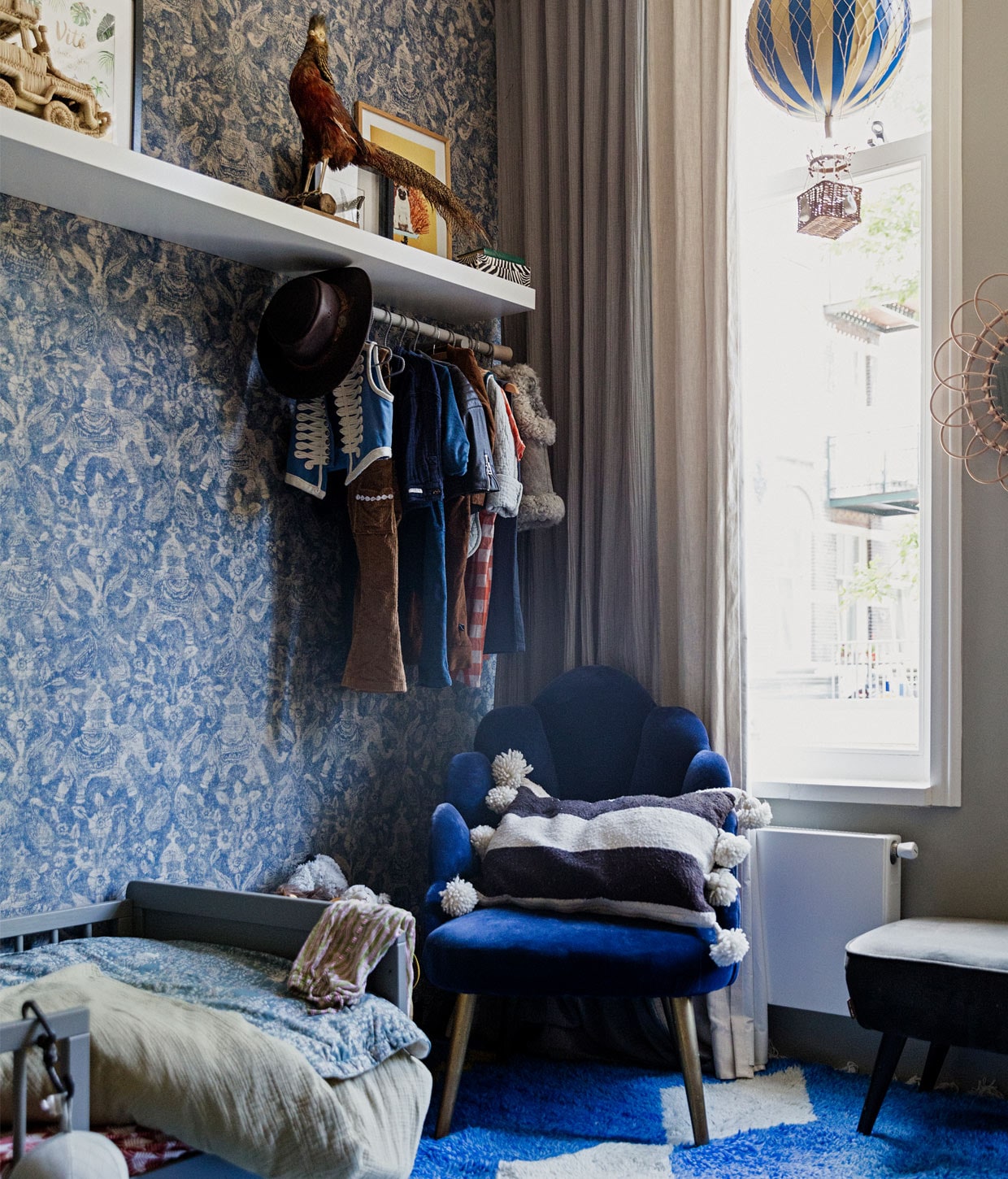 Behang Arte Walls, muurverf French Grey van Farrow &amp; Ball, raambekleding Numero 74, vloerkleed The Weavery, bed Petite Amélie, stoel Sissy-Boy, kussen in stoel The Souks, blauw beddengoed Garbo &amp; Friends, zachtgroen beddengoed Petit Sheets, rood beddengoed onbekend, wandplank Ikea, kunstwerk links naamposter van Fleur des Fleurs, kunstwerk rechts foto van hond Willem door Daan Brand. Opgezette vogel van de rommelmarkt, luchtballon Anna + Nina, spiegel bloem gekregen, bankje Karwei.