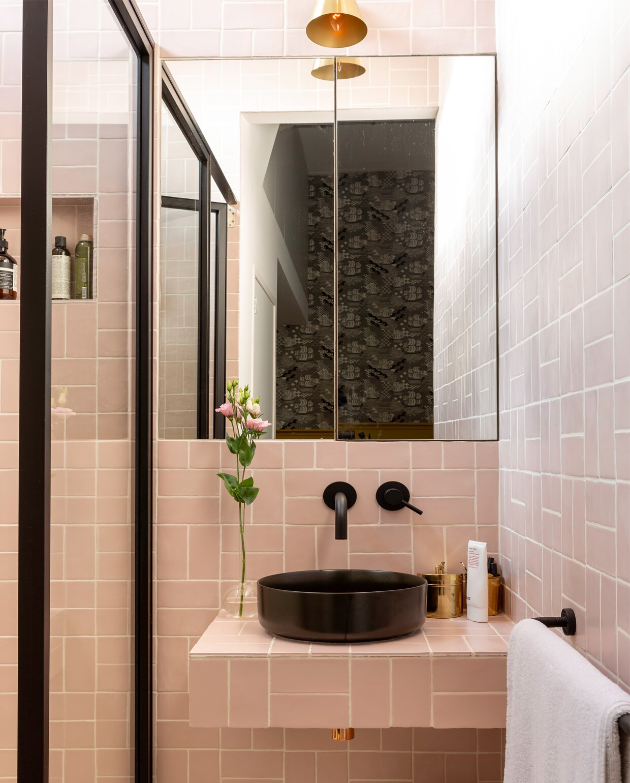 Op de muur rechthoekige tegels in poederroze, beide van Kol Tegels. Waskom M-Style serie 600 in 35,5×35,5 cm in mat zwart, kranen en siffon in brass, alles via Meijer Tegels &amp; Sanitair. Stalen douchedeur op maat gemaakt door Special Metal. Gouden lampje boven de spiegel van Soho Home.