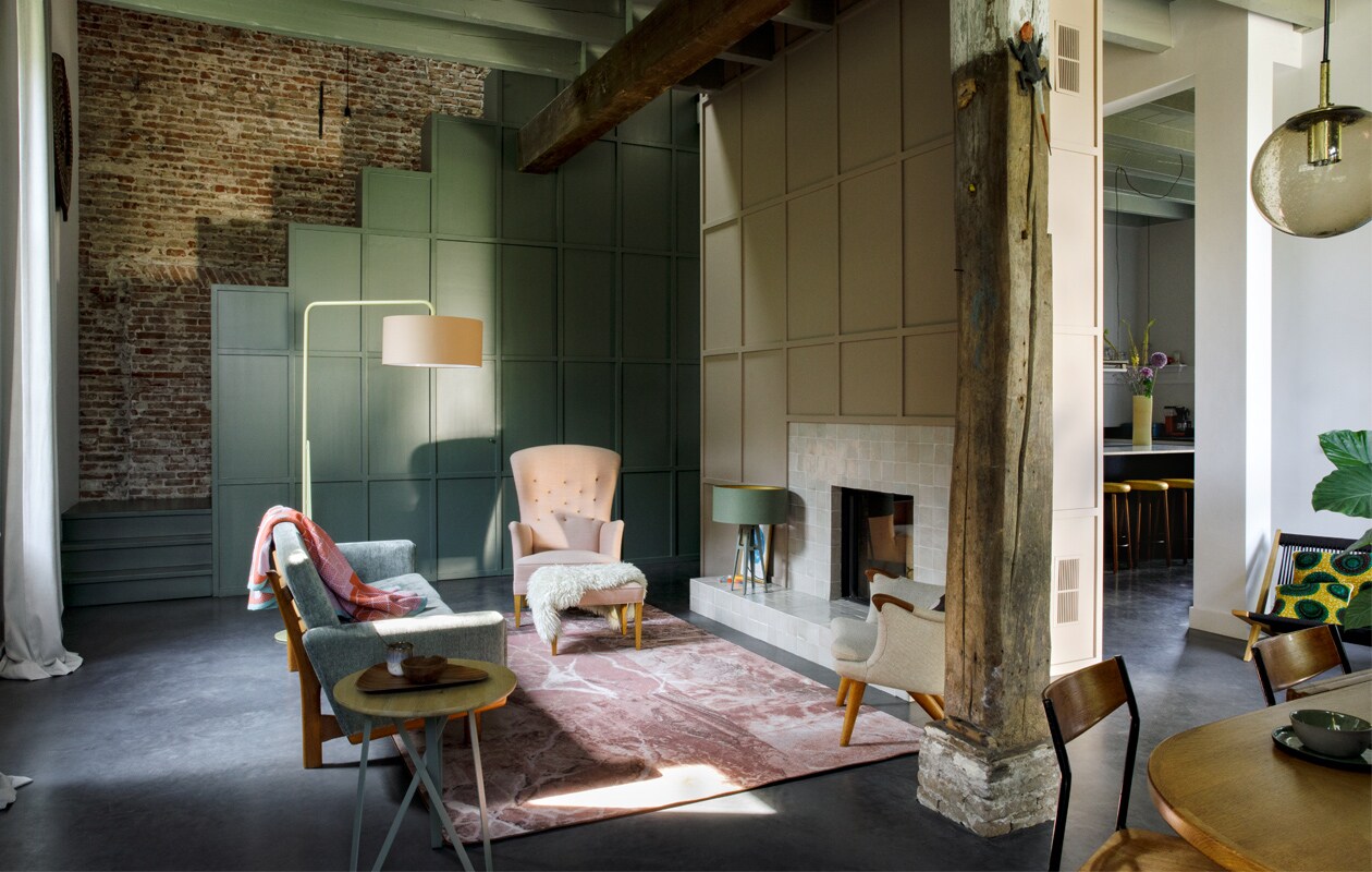 Gordijnen, vakken bij trap en vlakverdeling boven de haard eigen ontwerp. Verfkleur trap en trapkast Green Smoke, Farrow &amp; Ball. Tegels haard Rozetta. Vintage bank, fauteuils en voetenbank Morentz, plaid Sigrid Calon. Vloerkleed Sense of Marble van Desso. Lampen en bijzettafel Twister van Spoinq. Dienblad en kop herkomst onbekend. Schaal en schapenvacht lokale markt.