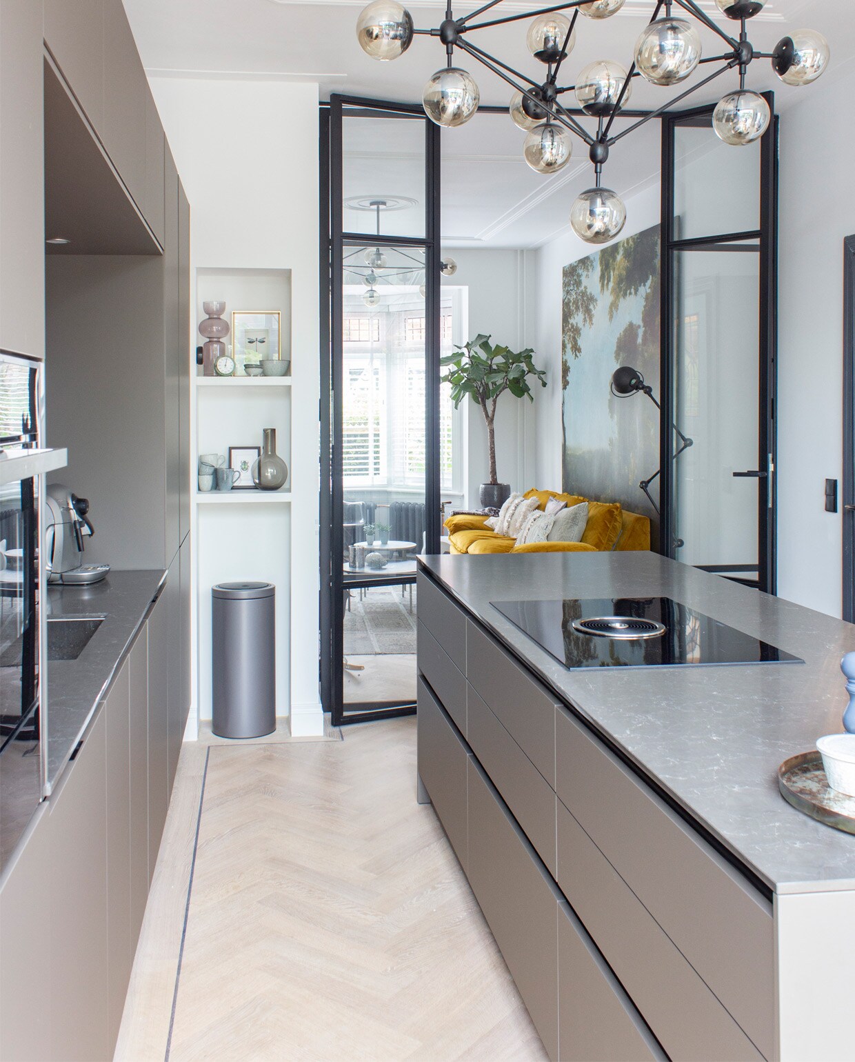 Siematic-keuken in kleur Umbra in matte uitvoering met een blad van composiet. Lamp met rookglas Loftlamp. In de nis: bruine vaas Karwei, bruine karaf H&amp;M home, mokken van Broste Copenhagen. De stalen binnenpui is gemaakt door Brink Stalen Deuren. Prullenbak Brabantia.