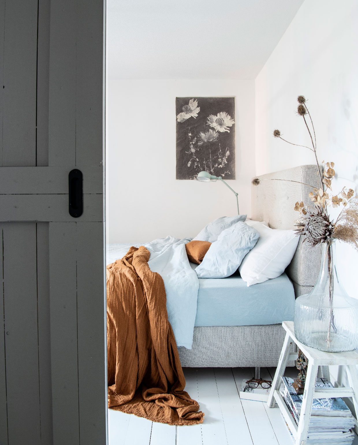 Houten vloer Pont Meyer Zaandam. Botanische print aan de muur Mo-ca. Bed Ikea. Lamp Made. Blauw beddengoed Dutch Home Fabrics. Plaid H&amp;M Home. Kruk tweedehands. Vaas op kruk Zuiver. Schuifdeuren gemaakt door aannemer.