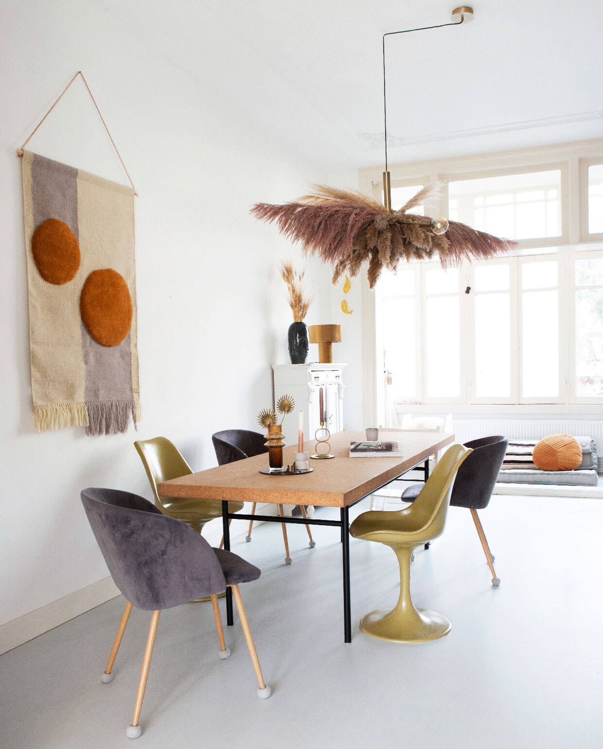 Kurken tafel Sinnerlig van Ikea, wandkleed Oyoy. Mosterdgele Tulip Chairs van Saarinen via Casa Gitane, grijze velours stoelen Søstrene Grene. Hanglamp van Frandsen via Loods 5, hangende pampasdecoratie zelfgemaakt door Susanne.