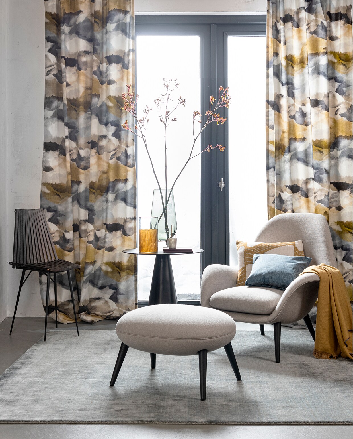 Gordijnen gemaakt van stof Minako in de kleur Gold/Charcoal van Harlequin, kussen van stof Kanako in de kleur Gold van Zoffany (stylelibrary.com). Houten stoel met metalen poten (paullelieveld.com). Bijzettafel van HKliving (vtwonen.nl). Vaas met deels amberkleurig glas, grijs kopje (loods5.nl). Blauwe vaas op messing poten van Menu, fauteuil van Fredericia, bijpassende poef, vloerkleed van Massimo (designtales.nl). Blauw kussen van denim Hardy black-out 35 (lifestyle-interior.nl). Okergele plaid (klevering.nl). Overig privébezit.
