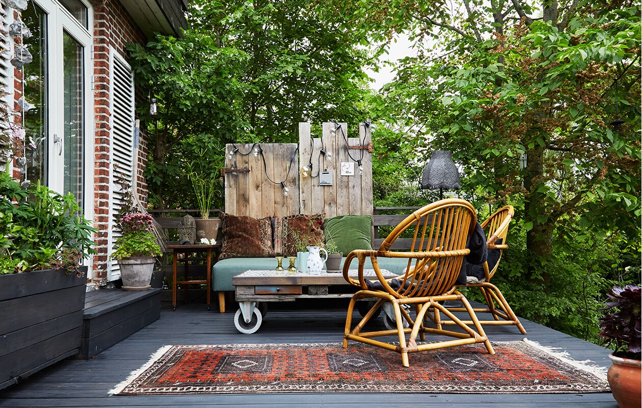 Het terras heeft verschillende niveaus en verbindt het huis met de tuin. Op het bovendek een oud loungebed bekleed met waterafstotende stof, een vintage rieten stoel en een zelfgemaakte pallet tafel op wielen met tegels van Arttiles. Erachter een paar oude deuren die een nieuw leven kregen als schutting. De lamp is van Ikea, de lichtketting kocht Trine bij de lokale bouwmarkt. Het grote tapijt vonden ze tijdens een reis naar Iran. De kussens zijn zelfgemaakt. Kleine lantaarn uit Marokko, tafeltje vintage. In de zelfgebouwde plantenbak staan lupines. Tegen de luiken klimt een clematis.