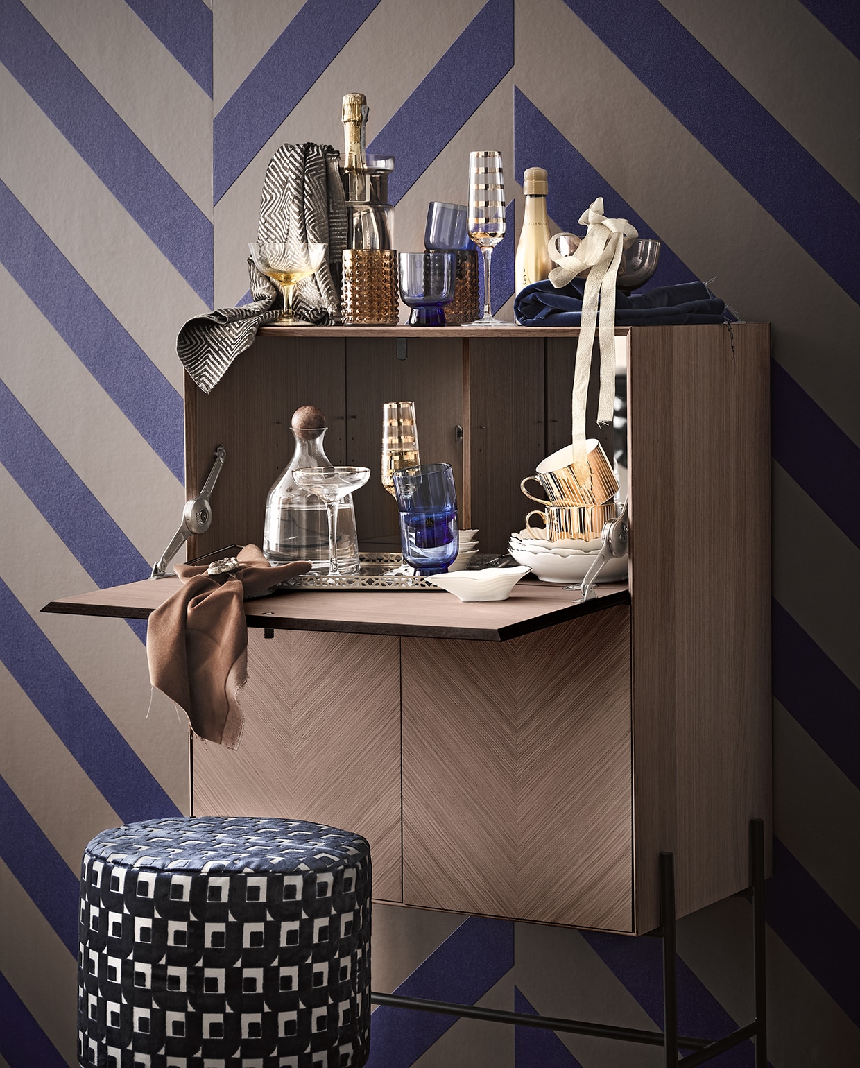 Behang tegen de muur Ulf Moritz Geometric 16404 (arte-international.com). Losse stoffen per meter: linksboven, van Nobilis, blauw velours rechtsboven van Designers Guild, reliëfvelours om poef (wva.nl). Bruine stof Colour Box Velvet f111/11045 (cole-and-son.com). Barkast Luxe met fineerafwerking (bolia.com). Kleine fles champagne, groot (bijenkorf.nl). Goudgestreepte kop-en-schotel, champagneglas met gouden streep, bruine mok, bruin glazen schaaltje, geel champagneglas, waterkaraf met houten dop, oestervormig schaaltje, witte borden, goudkleurig dienblad (hm.com). Vaas als wijnkoeler (loods5.nl). Overige privébezit.