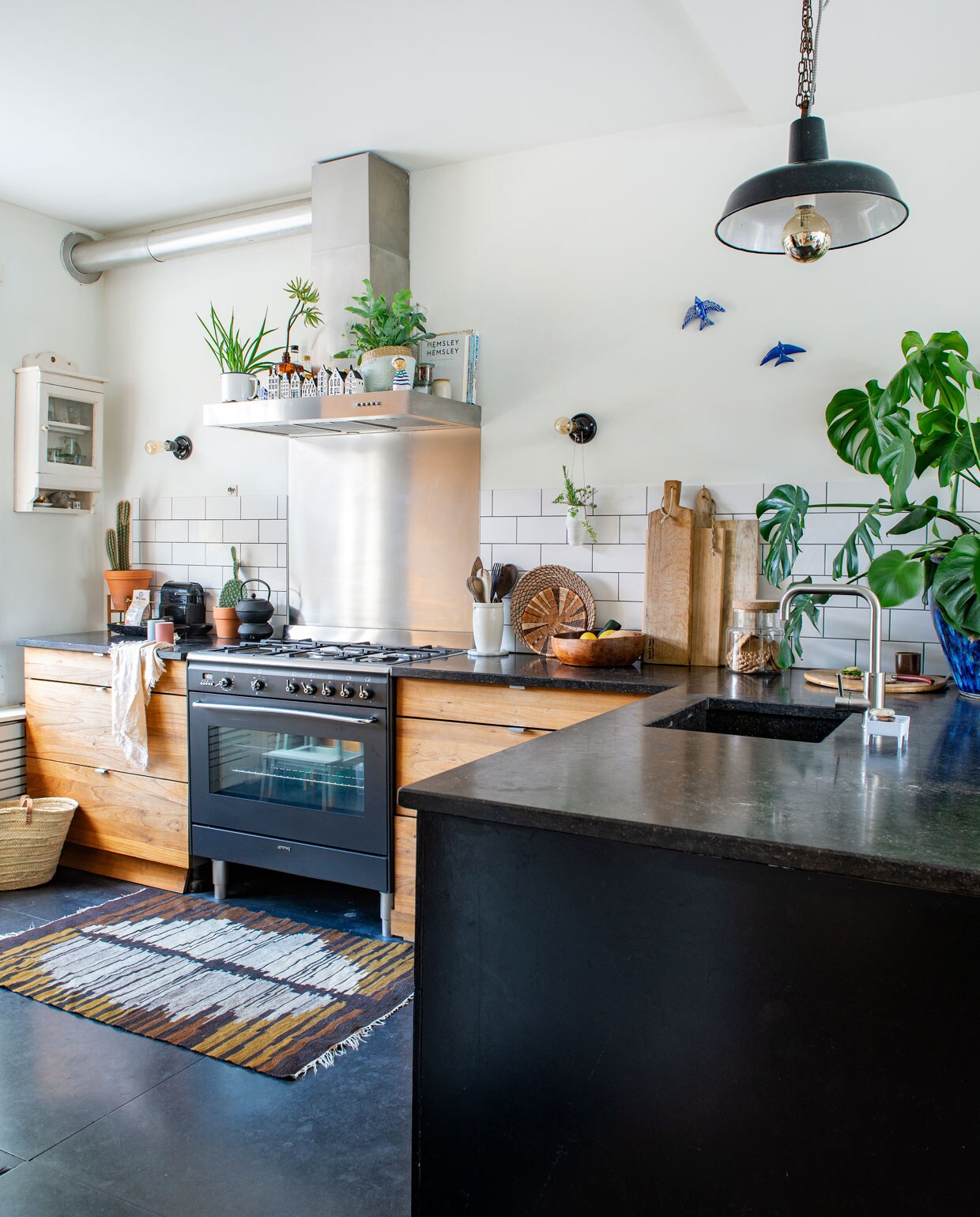 Keuken met op maat gemaakte fronten van iepenhout, werkblad en vloer van Belgisch hardsteen. Kraan Karwei, apparatuur Smeg. Hanglamp en vloerkleed Brilman Design. Wandlampen Bailey. Vitrinekastje Marktplaats, zeepbakje Foekje Fleur. Pot Ikea. Snijplanken Brood &amp; Plank, Ikea, Sissy-Boy. Lepelpot FairForward, onderzetters kringloop, vaasje aan muur Serax, vintage schaal cadeau gekregen, tas Etos, melkschuimer Nespresso, theepot en -licht Jang van Bredemeijer. Vogeltjes souvenir uit Portugal. Tegels Kol Haarlem, KLM-huisjes gespaard door Sanne.