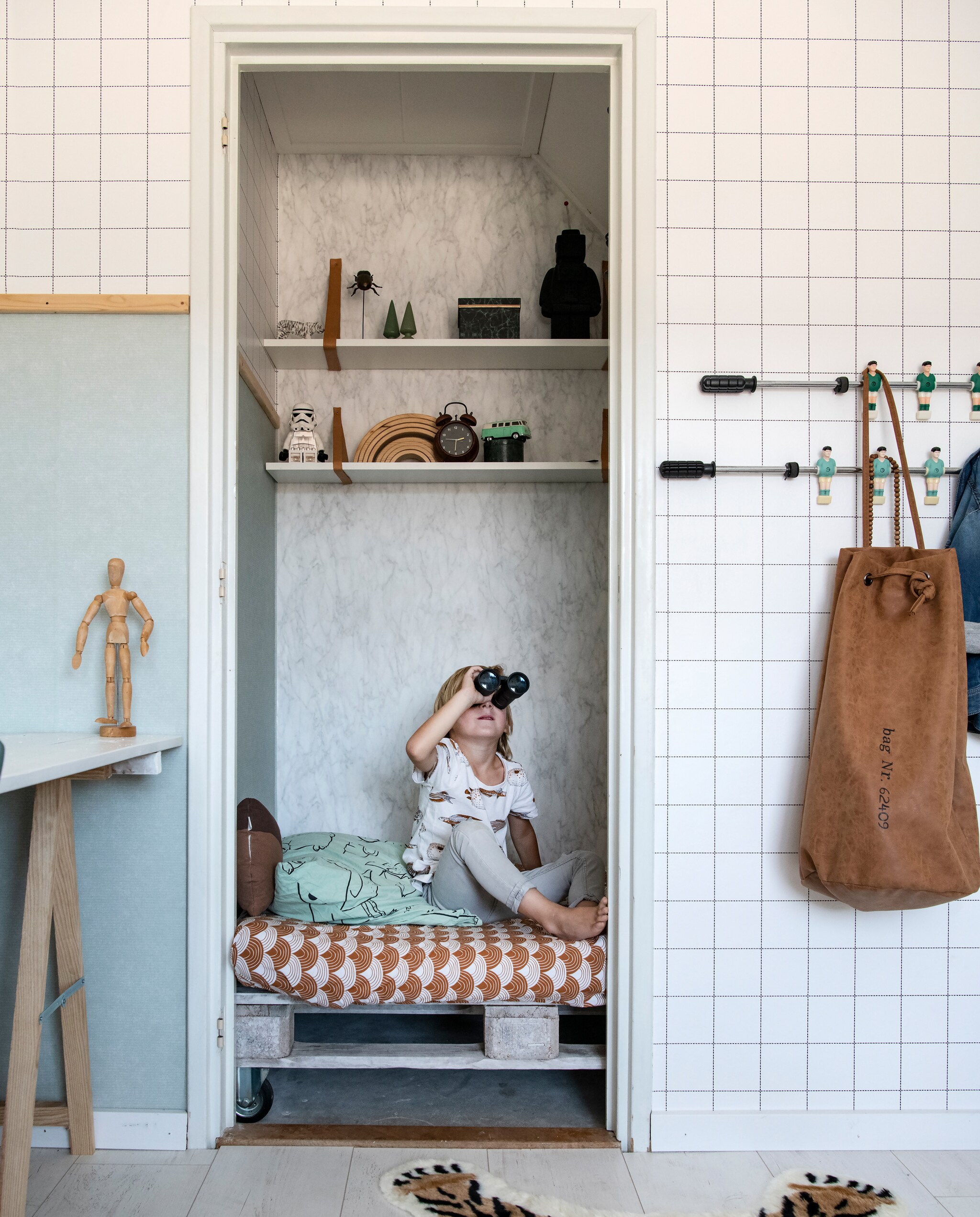 Lotte: ‘Na wat passen en meten bleek dat we in de inbouwkast op Mats kamer een soort bedstee konden creëren.’ Zijn bed is gemaakt van vlonders. Vloerkleed en marmerbehang van Kwantum. Tas van een tweedehands winkel. Beddengoed van Swedish Linens. De kapstok maakte Lotte van stangen uit een tafelvoetbalspel dat ze vond in een vuilcontainer.