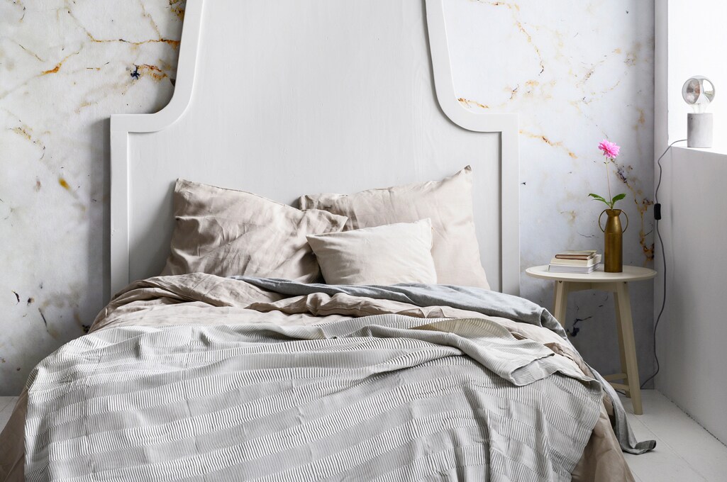 Fotobehang Milano met marmerprint, 300×300 cm, per rol € 149,- (karwei.nl). Bed-achterwand, 200×160 cm, prijs op aanvraag (com-anders.nl). Grijs-blauw plaid Vägmallä, 120×160 cm € 14,95, lichtbruin bedlinnen Puderviva, 240×220/60×70 cm € 79,95, bijzettafels Kragsta, 51×49 cm (hxØ) en 45×35 cm (hxØ) set van 2 € 79,95 (ikea.nl). Grijs flanellen bedlinnen vanaf € 17,50, hoeslaken Hotel vanaf € 18,- (hema.nl). Vaas van Bloomingville € 35,- (vtwonen.nl). Tafellamp Charis € 17 (kwantum.nl). Canvas Miss Susanna Gardiner, 25×30 cm € 45,- (werkaandemuur.nl). Overig privébezit.