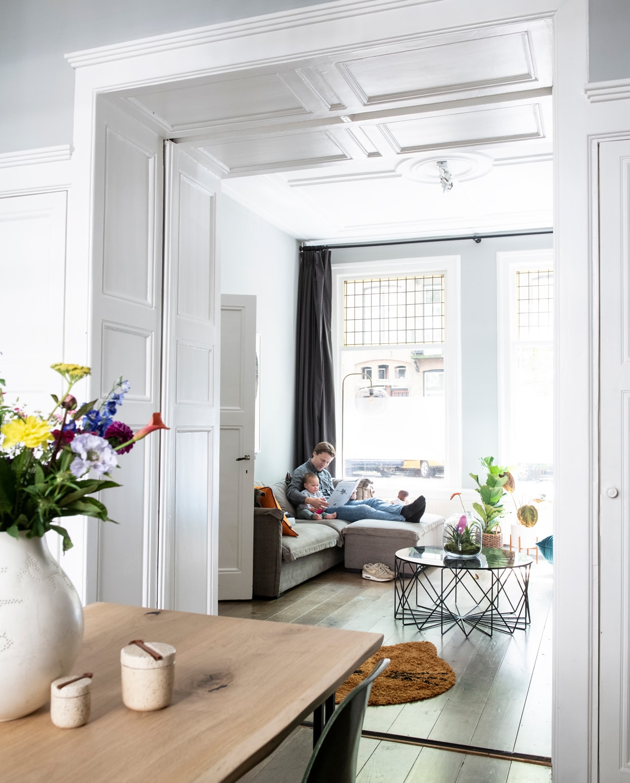 Alle oude details in het huis zijn origineel. De bank kochten Anne Mei en Hans nieuw bij een veiling. Erop liggen kussens met bloemen van Palais en kussens met bladprint van Sissy-Boy. Koffietafel en staande lamp met glazen kap van Bolia, op de eikenhouten vloer ligt kleed Cat van Liewood. Het kunstwerk aan de muur is door Anne Mei gemaakt. Plant en pot van Plantsome.