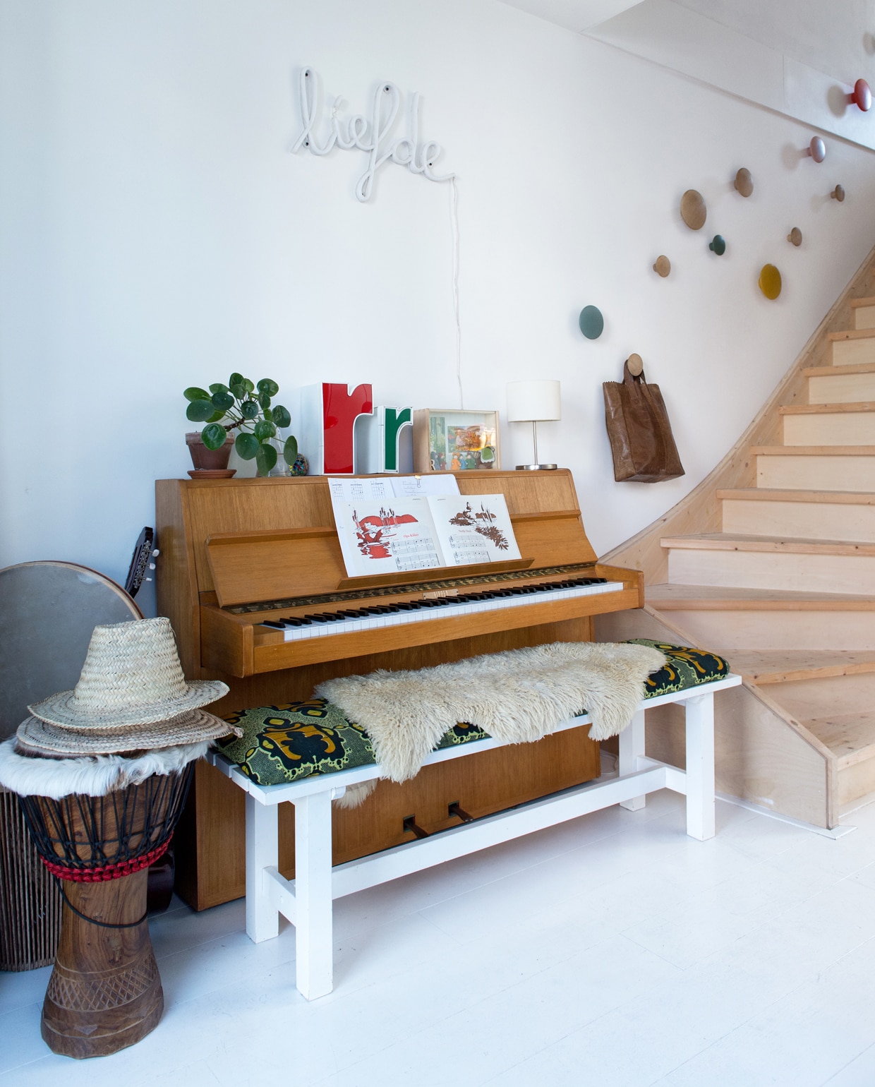 Tussen de voordeur en de trap staat een piano die van Michiels oma was. Bankje Ikea, kussen zelfgemaakt. Gekleurde doosletters Westside Den Bosch. Neon-lamp Liefde van Zilverblauw. Aan de muur The Dots van Muuto die dienstdoen als trapleuning.