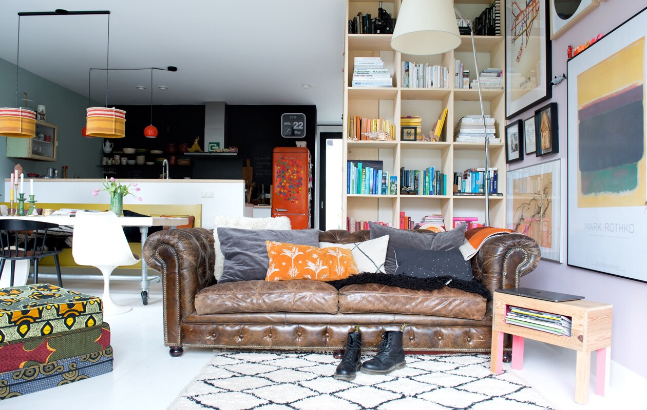 Rond het trapgat is een kast gebouwd voor keukenspullen en boeken. Jet en Michiel lieten hem maken bij dewinkeldiewerkt.nl. De vloer is gemaakt van duurzame eikenhouten duoplanken, gekocht bij Fairwood. Kunst van boven naar beneden: Tate Gallery By Tube poster gekocht via The London Transport Museum, foto Frida Kahlo, kunstwerkje gemaakt door de opa van Michiel, onder een eigen werk van Jet. Grijze kussens bank Madam Stoltz, oranje kussen The Pillow Room. Zwarte eetkamerstoel Hay.