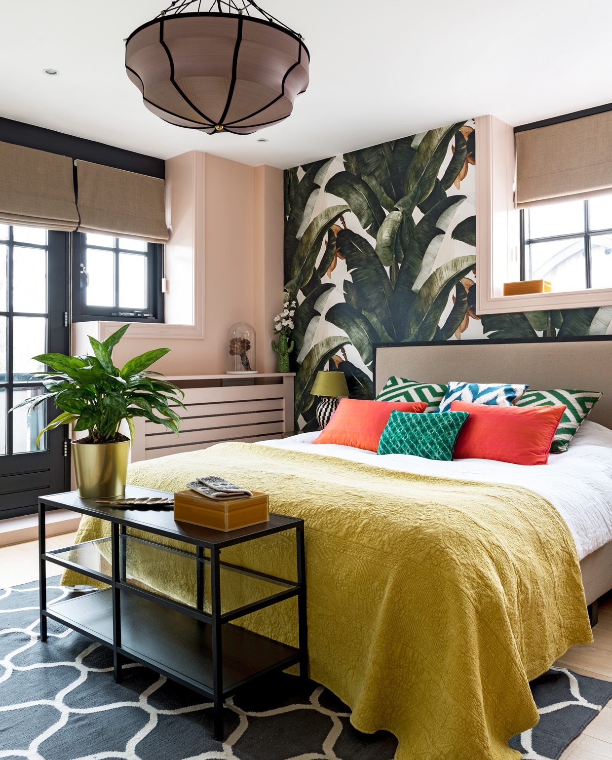 Behang van Lemon. Hanglamp van Gong. Headboard bed van Studio Sophie. Sprei van Zara Home. Groen kussen van Elitis, rode kussens van de groothandel en Ikat kussens van Eveline Interieur. Zwarte sidetable, vloerkleed en plantenpot Ikea. Glazen stolp met gedroogd bruidsboeket. Lamp met zwart-witte voet van Flamant. Roze verf Pink Ground van Farrow &amp; Ball.