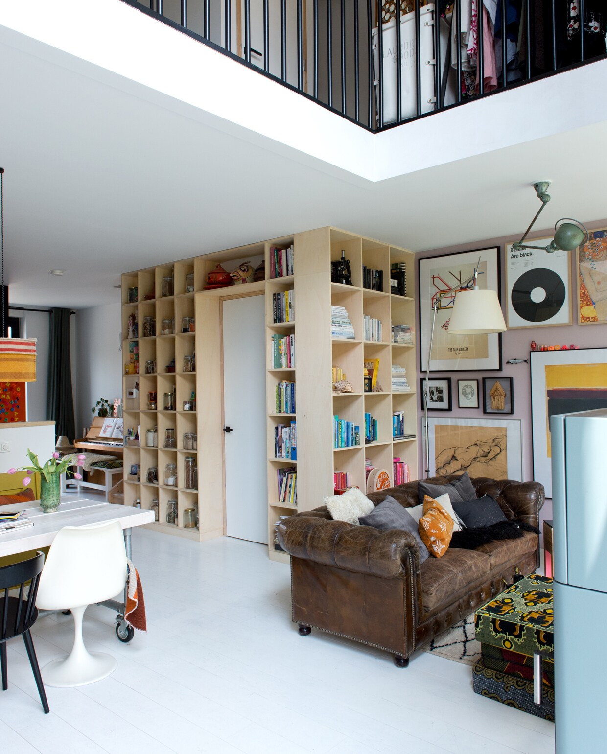 Rond het trapgat is een kast gebouwd voor keukenspullen en boeken. Jet en Michiel lieten hem maken bij dewinkeldiewerkt.nl. De vloer is gemaakt van duurzame eikenhouten duoplanken, gekocht bij Fairwood. Kunst van boven naar beneden: Tate Gallery By Tube poster gekocht via The London Transport Museum, foto Frida Kahlo, kunstwerkje gemaakt door de opa van Michiel, onder een eigen werk van Jet. Grijze kussens bank Madam Stoltz, oranje kussen The Pillow Room. Zwarte eetkamerstoel Hay.