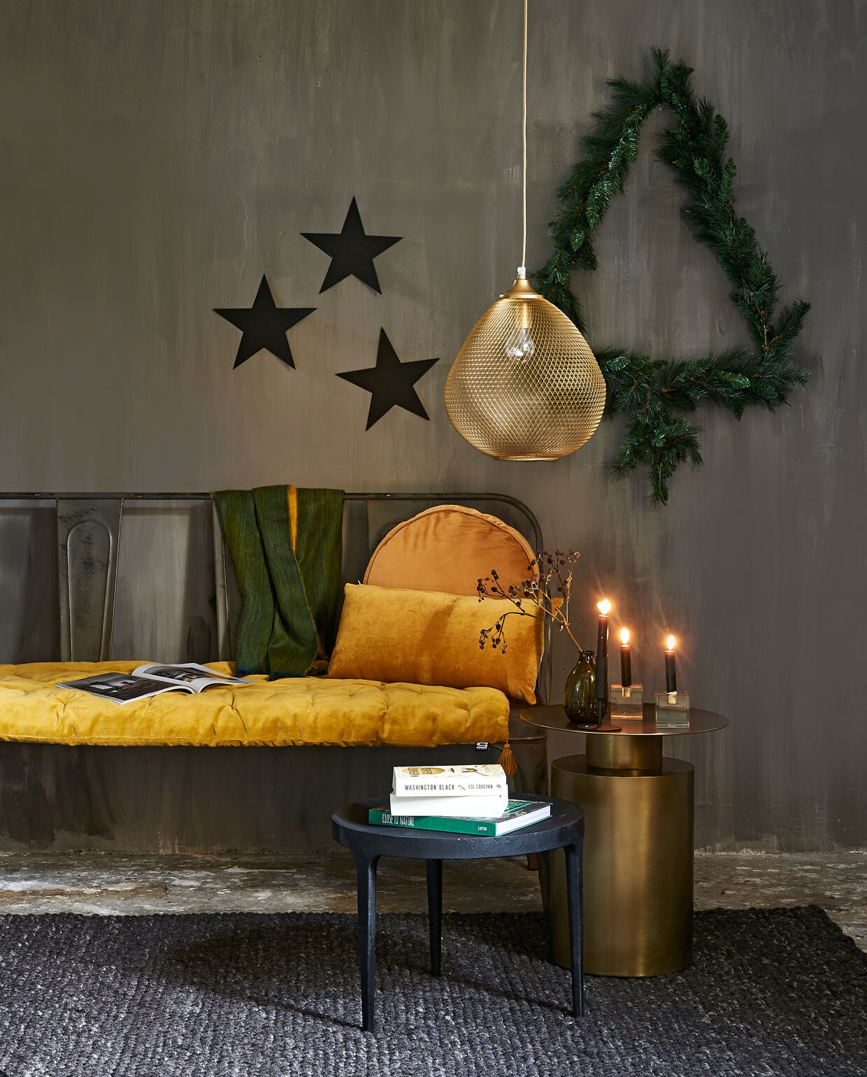 Metalen bank, groen vaasje (weldaad.com). Lamp (bodilson.com). Koperen tafel van Loods 5 Design, zwart tafeltje van Loods 5 Design (loods5.nl). Kerstslinger waarmee de kerstboom op de muur is gemaakt (rivieramaison.nl). Geel fluwelen kussen op bank, rechthoekig kussen, rond kussen, groen-gele sjaal (loods5.nl). Vloerkleed Hjorthede, 170×240 cm (ikea.be). Amberkleurige kristallen kandelaar vierkant (hkliving.nl). Sterren zelfgemaakt. Vaasje privébezit.