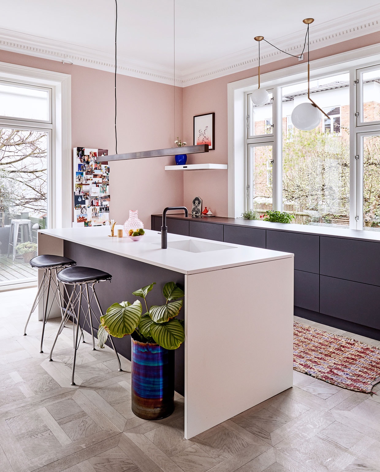 Bleekroze muren verrassen in de keuken, die wordt gedomineerd door strakke units gemaakt door het Deense bedrijf UnoForm. Het blad is van composiet. Werklicht wordt voorzien door een handgemaakte donker koperen lamp van Anour, de bollampen IC – met koperen armatuur – zijn van Flos. De krukken zijn van Overgaard &amp; Dyrman, de geglazuurde vaas is een ontwerp van Tom Dixon voor Roomstore. Op de vloer ligt een Boucherouite, een traditioneel, handgeknoopt kleed uit Marokko.
