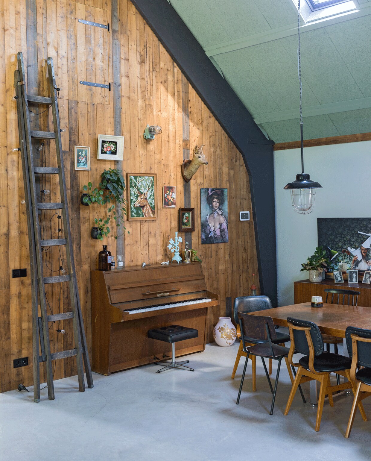 De houten muren zijn gemaakt van een partij verweerd dakbeschot. De ladder tikte Wende op de kop via Facebook, het lichtsnoer eraan is van Intratuin. Ljstjes aan de wand via brocantemarkten in Frankrijk. De piano is van een collega geweest, erop flessen van Davidowski, kandelaar Rice. Tafel van Mooi Muf, eromheen leren stoelen van Montmartre, een brocantemarkt in Gouda, stalen stoel Davidowski, spijlenstoel Tony’s Garage Sale. Grote foto op het dressoir van Hugo. Hanglamp Vroeger &amp; Toen.