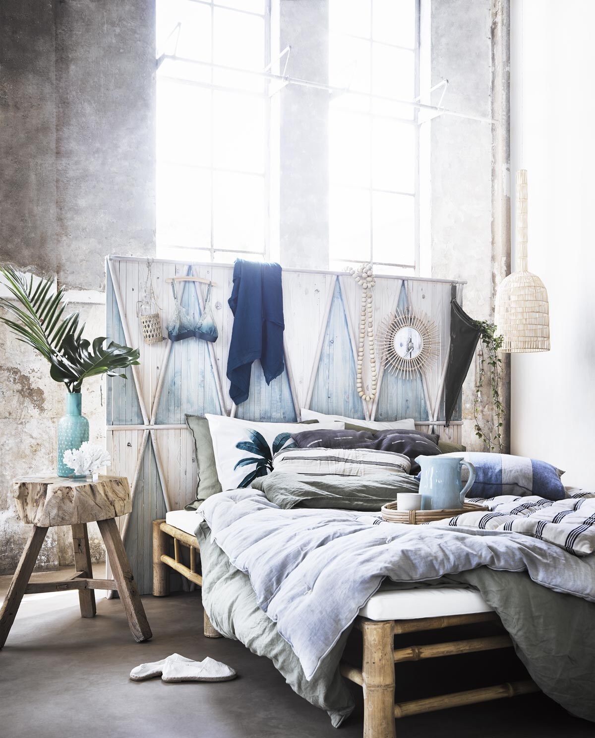Novilon vloer Zand (vtwonen.be). Muurverf Ibiza op muur rechts (bauwerk.com.au). Daybed van Tine K Home (wehkamp.nl). Hoofdbord van blank hout, groene plantenzak, lichtblauwe kan (com-anders.nl). Oud houten tafeltje, zeeblauwe sjaal, ketting met blank houten kralen (sukha.nl). Hanglamp (couleurlocale.eu). Kunstkoraal op voet (zarahome.com). Witte zomerschoenen, groen met wit dekbedovertrek en kussenslopen met palmprint, blank houten spiegel, rond rotan dienblad, witte mok (hm.com). Glazen fles met gevlochten touw, groen dekbedovertrek met twee kussenslopen (loods5.nl). Blauwe beha, blauwe vaas (maisonnl.com). Blauw en grijs kussen met witte streepjes, lichtblauwe plaid, witte plaid met blauwe streep (letoile.store). Stof van blauwe kussen met beige streep (wva.nl).