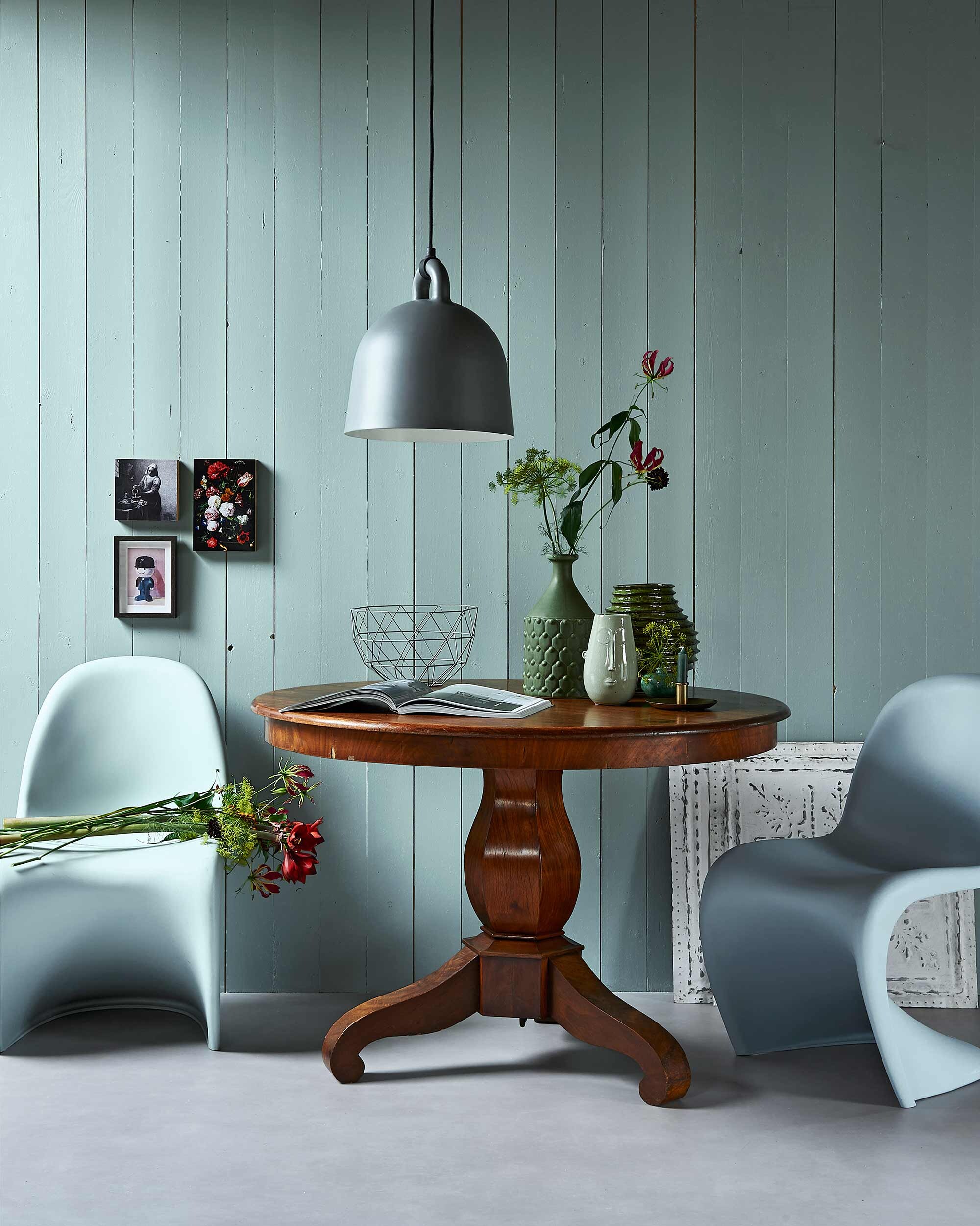 Vloer kiezel (Novilon van ). Wand geschilderd met muurverf Classico in kleur Polar Blue (). Antieke tafel (). Hanglamp Bell, stoelen Panton Chair, hoge groene vaas, vaas met gezichtje, klassieke print op houten blok (). Groen geribbelde vaas, koperen kandelaar, metalen wandplaten ().