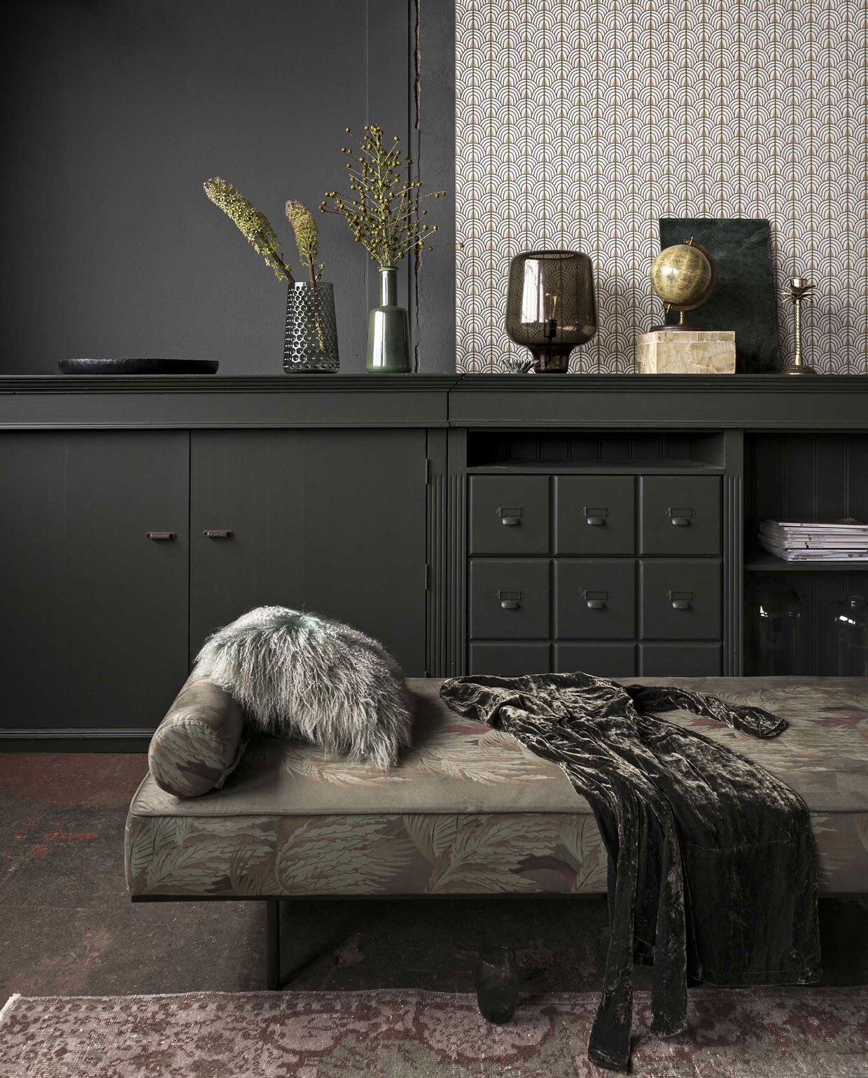Op de muur muurverf kleur Scree van Little Greene, behang Coordonnee Core collection 6600045 (). Daybed bekleed met fluweel (). Harig kussen Tibet Jade, glas (). Donkergroene kast (). Vloerkleed (). Zwart dienblad, glazen vaas met reliëf van Bloomingville, glazen stolp rechts in kast van Bloomingville, glazen stolp links in kast van Casa Vivante (). Groene glazen vaas (). Bloemobject van keramiek (). Lamp van Northern Lighting (). Wereldbol, lichtgroen doosje van in mozaïek gelegde stenen, messing kandelaar (). Groen marmeren blad van Madam Stoltz (). Fluwelen ochtendjas ().