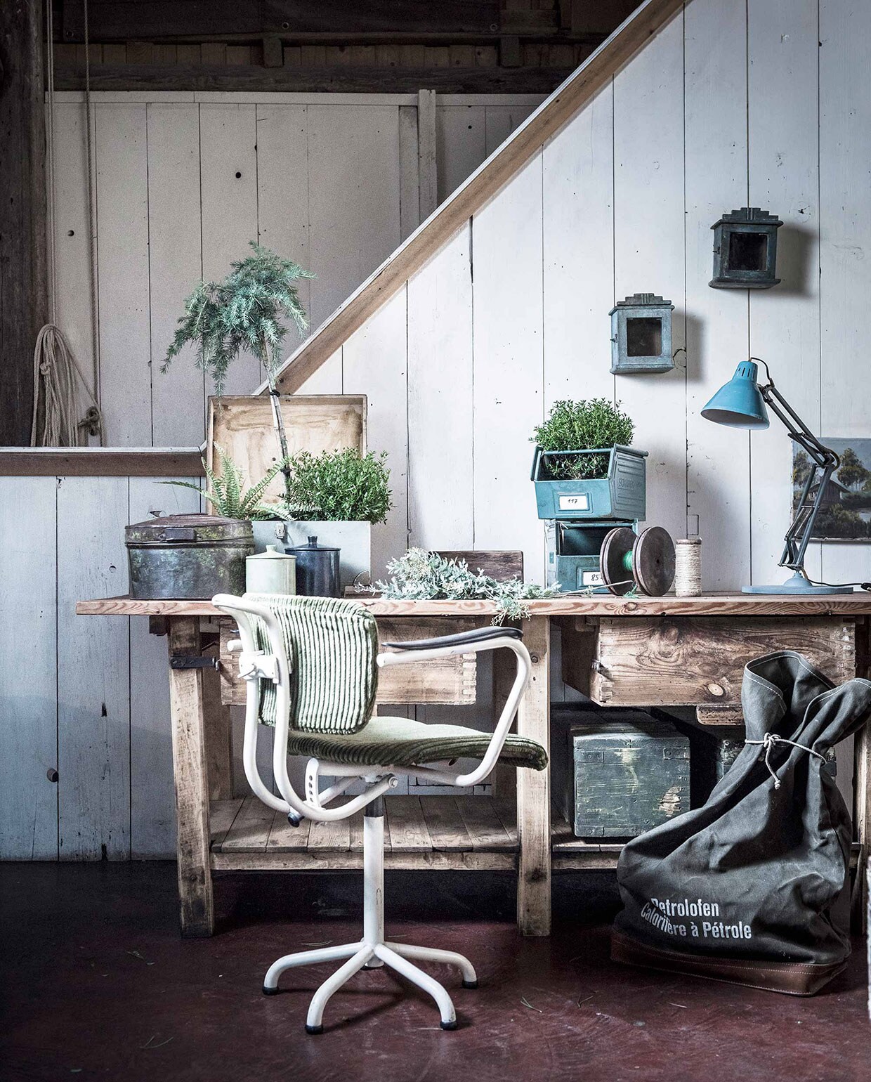 Houten werkbank, mintgroene kist, schilderij, legerkisten (). Bureaustoel, bureaulamp (). Ovale stalen kist, kastjes aan de muur (). Zwarte pot, witte pot (). Stalen opbergbakken, garenhouder met groen garen (). Klos met wit touw (). Groene legertas ().,