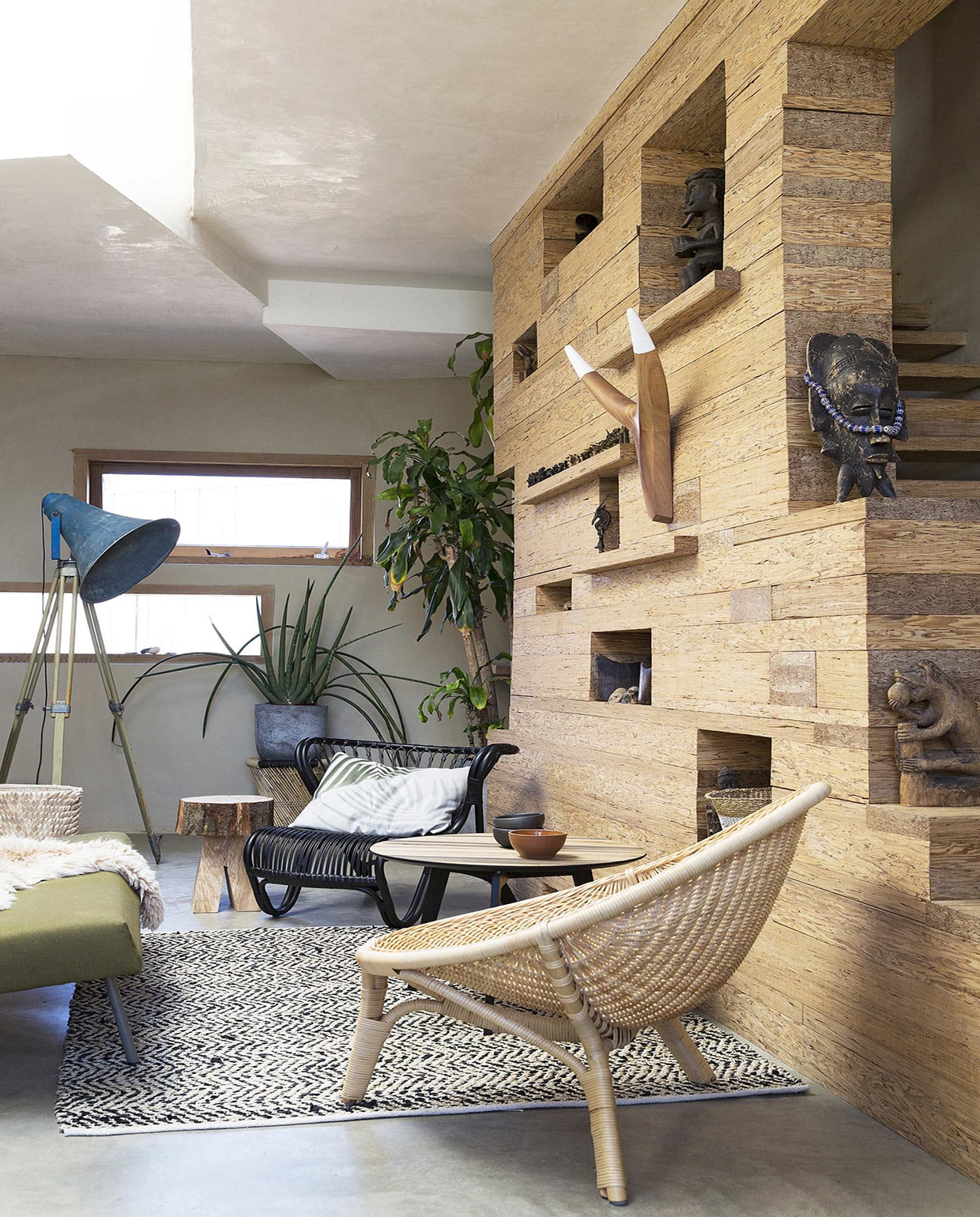 Wand langs trap, ontworpen door Nicky Zwaan en Joris Brouwers/House of Bouw, gemaakt van PSL (Parallel Strand Lumber) dat in de VS wordt gebruikt als constructiemateriaal in de huizenbouw, hier niet te koop (House of Bouw). Vloerkleed met zigzagpatroon van leer en katoen, gevlochten krukje Mudda Stool onder plant (Zenza Home). Stoel in voorgrond Rana Chair NO-13-CU naturel, stoel Viggo Boesen zwart V8-11-PS, eiken salontafel Slo Savon (Design 020). Kussen met palmbladprint (Store Without a Home). Mand Porcupine naast zetel, nguni hoorns in wand (From Africa With Love). Kommen zwart en rood van Beate Snuka (Frozen Fountain). Staande lamp (Raw Materials). Blokkrukje (Neef Louis). Grijze pot (Tuincentrum Osdorp). Overig privébezit.