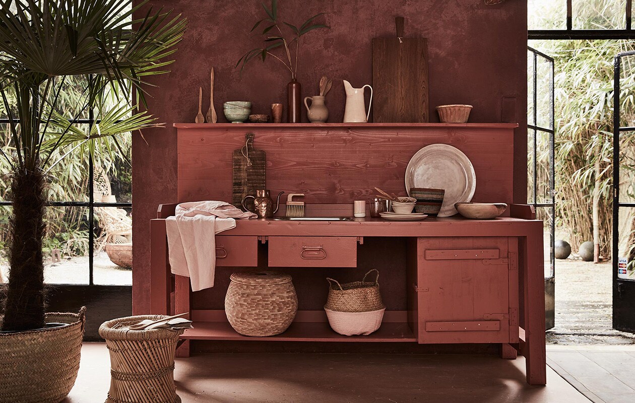 Keuken met werkblad (Combitex). Mand (met palm erin), mand met deksel, linnen schort, houten schaal, houten lepels, gestreepte mand rechts op het keukenblad (Couleur Locale). Mand met roze bodem (Sissy-Boy). Houten planken, grote lila schaal, hoge kan, kommen (op werkblad), borden, linnen theedoek (Meker Wonen). Puddingvorm (De Weldaad). Koperen kan (All the Luck in the World). Roze beker (Six and Sons). Groene kommen (Serax). Houten bakje op plank (HK Living). Houten beker (Raw Materials). Bamboe kruk (Zenza). Bruine oude kan privébezit.[/caption]