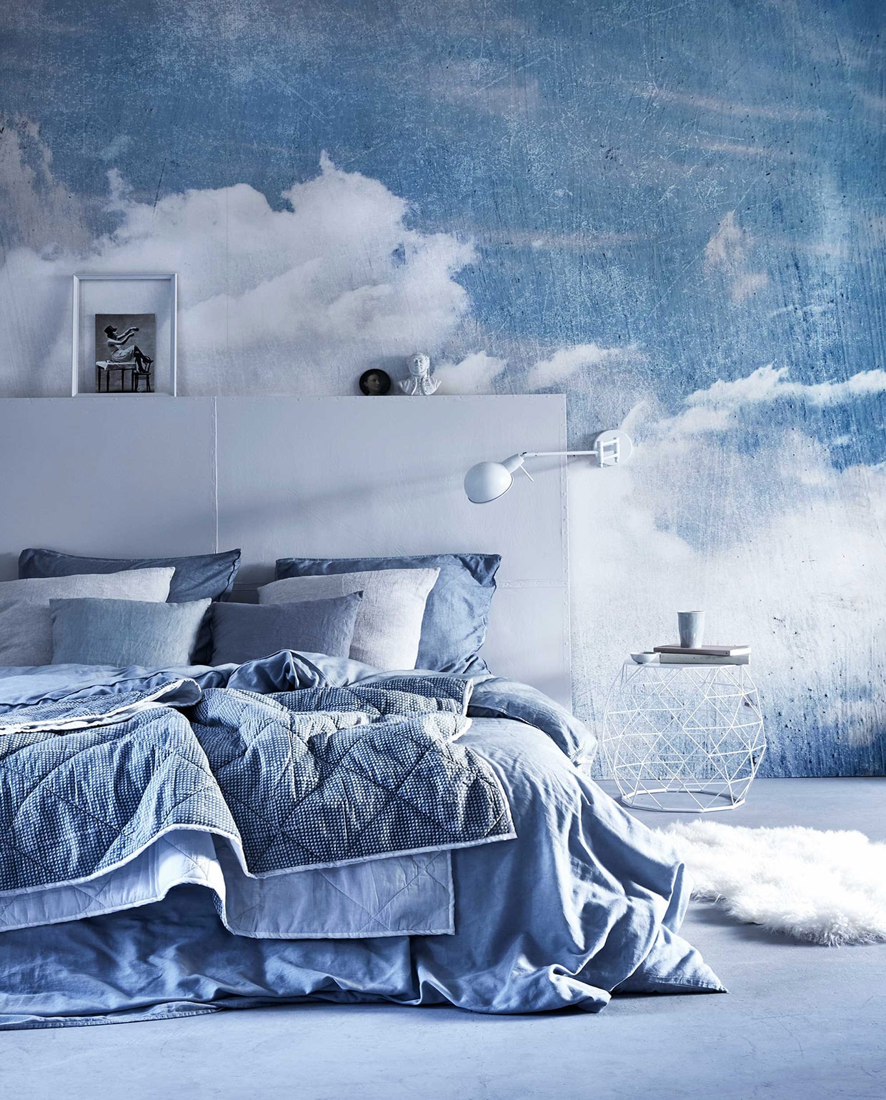Cloud Puff van Rebel Walls (). Hoofdbord (). Beddengoed van satijnkatoen (). Beeldje (). Geprint plaid, kleine blauwe linnen kussenhoezen, schapenvacht (). Linnen  in Misty Grey (By Mölle). Bijzettafel, wandlamp Glasgow (). Onderzetter (). Fotolijst en boeken privébezit.