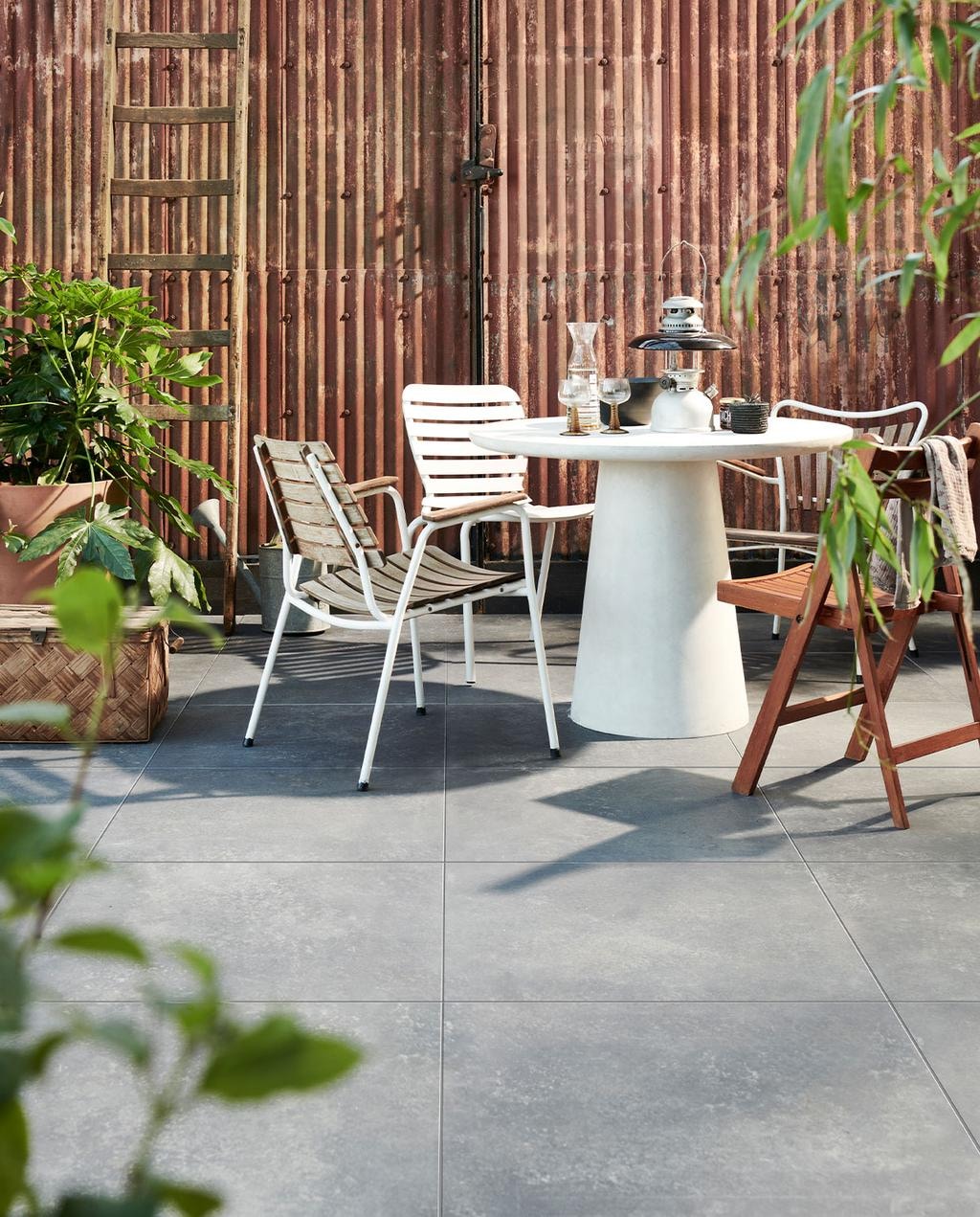 Buitentegels Solostone Uni Chateau Black, 70x70x3,2 cm per m² € 69,95. M.m.v. basiclabel.nl (betonlook tafel van Damon), vtwonen.nl (witte metalen stoel van Zuiver), com-anders.nl (oude ladder, houten kist, oude gieter, klapstoel, lantaarn, rieten mand), deens.nl (bloempot met onderstel van Ferm Living, stof over stoelleuning, kan, glazen), vandijkenko.nl (stoelen van ijzer en hout).