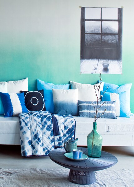 Behang Saraille Aqua van Designers Guild per rol € 242,-, blauwe en turkooizen kussens met witte baan Savine van Designers Guild € 60,30, de grote kussens zijn gemaakt van stof Padua Aqua van Designers Guild € 106,70 p/m (Wilhelmine van Aerssen). Guilt € 109,95, grijs-wit dip dye kussen € 40,-, langwerpig grijs-wit tie dye kussen € 40,-, kussen indigo met cirkels € 40,- (Bloomingville).