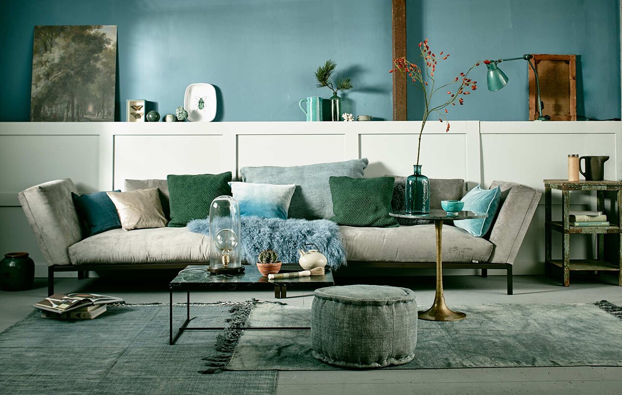 Muurverf Pure &amp; Original Classico krijtverf, kleur Libo Bay per liter, lak op lambrisering Pure &amp; Original Classico Oil Eggshell in de kleur Silver Clay, (De Ru). Long chairs City, blauw linnen kussen van Nordal (vtwonen.nl). Ecru kussenhoes van fluweel, groene gebreide hoes, blauwe fluwelen hoes met kleurverloop (H&amp;M). Groot kelimkussen (Sukha Amsterdam). Schapenvacht (Nordal). Lichtblauw kussen (De Weldaad). Vloerkleed links, ronde poef, vloerkleed rechts (Aai made with love), vaas links van zetel (Raw Materials). Salontafel met groen marmeren blad en stalen frame, stolplamp (Serax). Ovaal zwart dienblad (Maison NL). Theepot (Meker Wonen). Marmeren kwast (Zenza). Hoge, ronde bijzettafel met bronzen voet (Madam Stoltz). Glazen vaas (H&amp;M). Kom (In Petto). Verweerd tafeltje rechts (Raw Materials). Kartonnen koker (Neef Louis). Kan (Ikea). Op de lambrisering, v.l.n.r. afbeelding van schilderij via het Rijksmuseum, geprint op karton vanaf (Repros), marmeren bol (Six and Sons), potje (kringloopwinkel), groene steen op voet (Fleurmonde), schaal met kever (Maison NL). Midden: Portugese kan (In Petto), groene fles (Brût Amsterdam), koraal (Fleurmonde), grijs kopje (Ferm Living). Rechts: groene atelierlamp Wielum (Brût Amsterdam), schilderdoek, omgedraaid (Six and Sons). Overig privébezit.