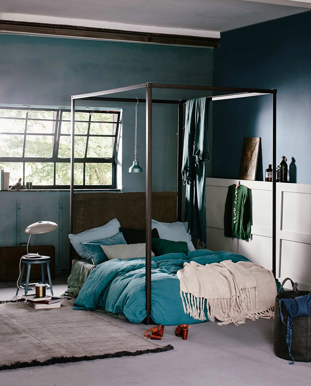 Hemelbed, op maat gemaakt (Combitex). Op bed: linnen kussensloop, linnen dekbedovertrek, 220×240 cm, linnen laken (Couleur Locale). Zeeblauw fluwelen kussen (De Weldaad). Twee groene kussenhoezen, gebreide beige plaid (H&amp;M). Kussen in lichtgroen canvas (Nordal). Hangende sprei (Hammam34). Hanglamp (Brût Amsterdam). Kruk (Madam Stoltz). Vintage leeslamp, schilderij op lambrisering, mand rechts vooraan (Six and Sons). Koffer, duif (De Weldaad). Groene gemarmerde beker (H&amp;M). Vloerkleed (Aai made with love). De muur bij het raam is geschilderd met muurverf Pure &amp; Original Classico krijtverf Blue Reef en de zijmuur in Libo Bay, lak op lambrisering van Pure &amp; Original Classico Oil Eggshell, kleur Silver Clay per liter (De Ru). Overig privébezit.