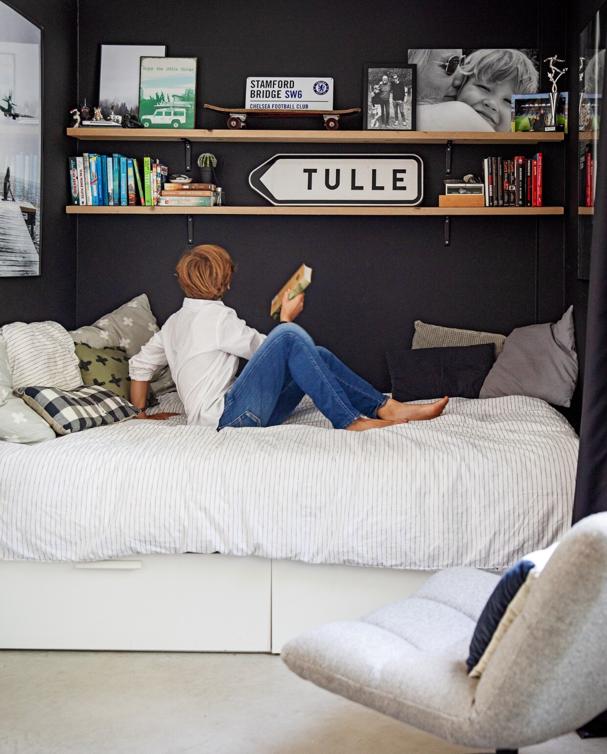 Vloer gevlinderd beton van Bull beton, vloerkleed HKliving. Muurkleur zwart Ral 9005, wandplanken oude steigerplanken Berko. Op de planken: ijzeren borden met afbeeldingen Loods 5, Stanford bridge uit Londen, privéfoto’s, voetbalprijs, verkeersbord Frankrijk. Bed Ikea, beddengoed Vandyck. Kussens (v.l.n.r.) plusjes zelfgemaakt, blokjes Loods 5, groene plusjes Intratuin. Stoel Dutchbone, kussen Deens.