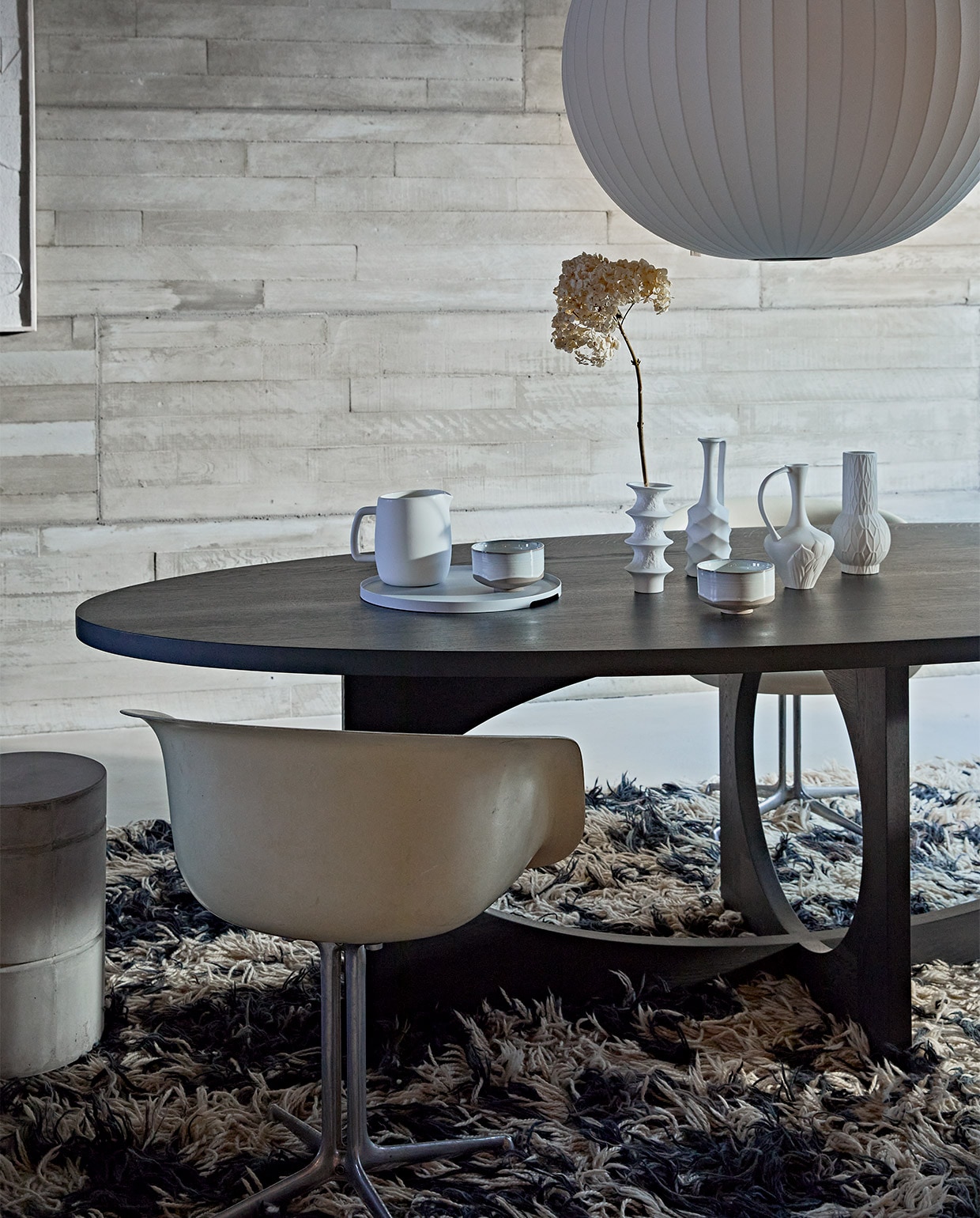 Zwartgebeitste massief eikenhouten ovale tafel van ontwerper Sarah Trenite, 260x115x75 cm (lxbxh), prijs op aanvraag (arpdesign.com). Vintage La Fonda kuipstoel van Eames voor Herman Miller, prijs op aanvraag (pietjonker). Grote witte vazen Cobra Bubble tall € 120,- en Double Bubble tall € 135,- van 101CPH (edha-interieur). Schilderij met reliëf, 100×70 cm € 199,- (urbannatureculture). Kleine vazen van HKliving, set van 4 € 129,95, keramieken waterkan van ontwerper Vincent van Duysen voor Serax, 1,5 liter € 25,-, hanglamp Nelson Ball Bubble van Hay, 68 cm (Ø) € 607,99, dienblad van Zone Denmark, 35 cm (Ø) € 50,-, kommen Merci n°1 van Serax, 12,5 cm (Ø) per stuk € 16,65 (vtwonen.be). Betonnen kruk van Marie Michielssen, 30×45,5 cm (Øxh) € 103,- (serax.com). Gedroogde hortensia’s (edenique). Soortgelijk vintage vloerkleed (vintagehoarder.com).