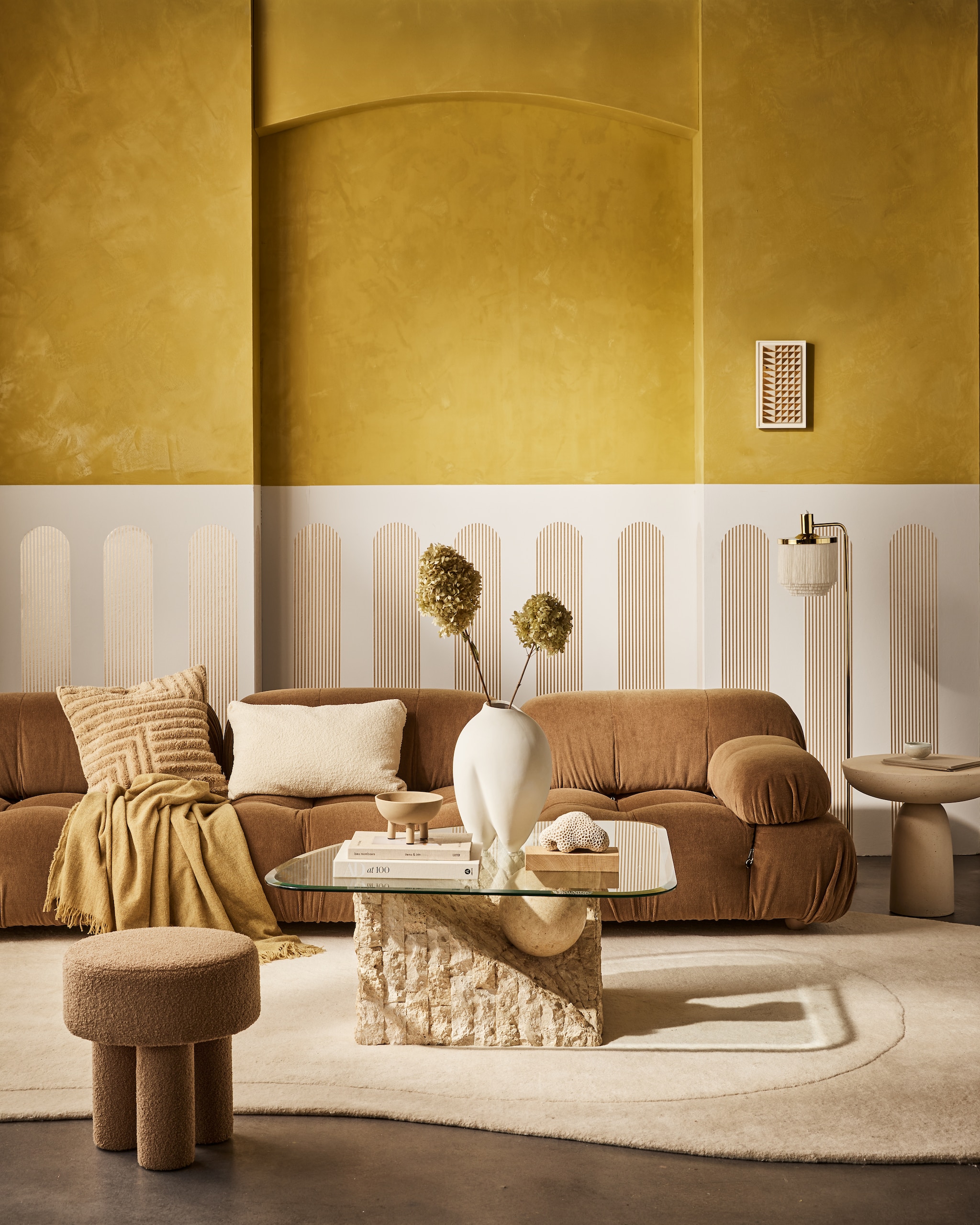 vtwonen collectie behangstrook Streep Gold-Egg White, 17,6 cm (b), rol van 10 m € 12,99. M.m.v. homestede.nl (bank, kussens, plaid), edha-interieur.nl (poef, bijzettafel), goeds.nl (vloerkleed van Layered, lamp), pietjonker.nl (salontafel), rollmannceramics.com (kunsttegel), lauthentique.nl (gele muurverf kalkverf in Korengoud 33, witte muurverf krijtverf in Krijt 63), 101cph.com (vaas, schaal). Overig privébezit. productie Tanja Saarberg | styling Cleo Scheulderman | fotografie Jeroen van der Spek | assistentie Kelly van Gendt | studio en decorbouw Studio Milo