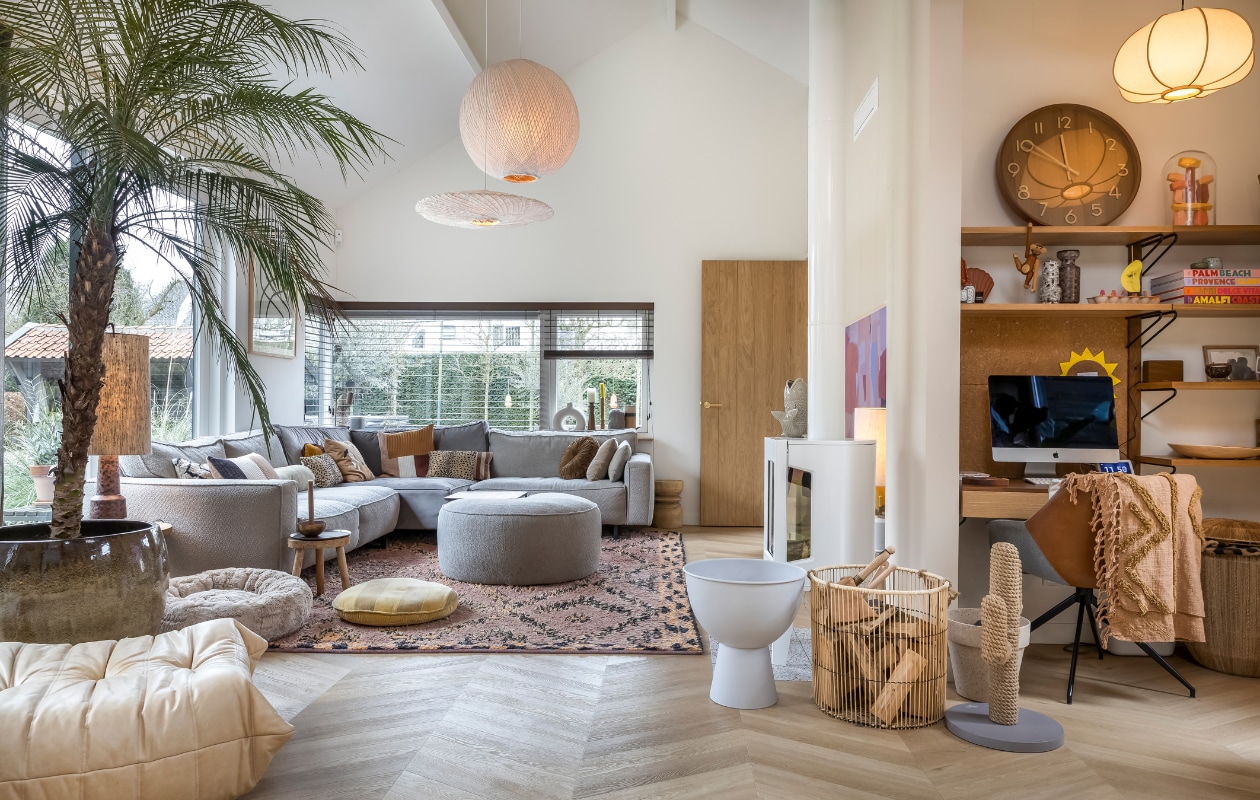 Muurverf Mushroom van Little Greene, verf plafond RAL 9010, pvc-vloer Therdex Chevron 6541. Hanglamp rond en ovaal HKliving. Kandelaars in vensterbank van links naar rechts: HKliving, Loft, grijze vaas onbekend, HKliving. Bank Melchior Interieur, kussens op bank: Ferm Living en HKliving. Kruk rechts van bank Karwei. Hocker grijs Melchior Interieur. Vierkant tafeltje, rond tafeltje, vloerkleed en geel kussen HKliving. Hondenmand dierenwinkel. Lamp op tafeltje HKliving. Plantenpot Jasmijn Bloembinders, palmboom Green Bubble. Togo fauteuil Ligne Roset. TV Samsung The Frame. Houtkachel Jotul, vis op kachel H&amp;M, lamp achter kachel en witte plantenbak HKliving. Mand Karwei. Krabcactus Designed by Lotte. Bloempot Karwei. Wandkast Made (niet meer verkrijgbaar). Klok Karlsson. Stenen onder stolp interieurwinkel in Los Angeles. Aapje Kay Bojesen. Vaasjes HKliving. Stoel Zuiver, plaid op stoel HKliving. Mand rechts van stoel Loods 5. Hanglamp H&amp;M. Raambekleding Onel Window Dressings. Overig onbekend. vtwonen 02-2023 | styling Karin van Schie | fotografie Henny van Belkom