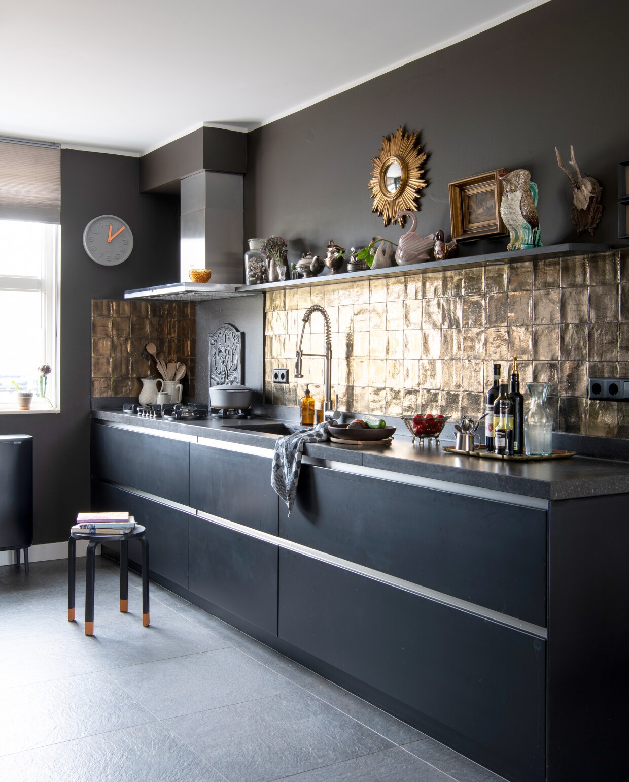 Muurverf Fading Horizon van Flexa, keuken zwart laten spuiten door Spuiterij Rood, gouden wandtegels Sanitairkamer, achterplaat fornuis zelfgemaakt van smeedijzeren haardplaat, klok Zuiver, kruk Ikea, vintage dienbladen erfstuk en rommelmarkt, keukenrek Ikea, slabestek antiek, vleesvorken en keukenmessen rommelmarkt, slabestek antiek. Overig onbekend. vtwonen 01-2023 | Productie Inside Homepage | Fotografie Louis Lemaire | Styling Anja Bas Backer