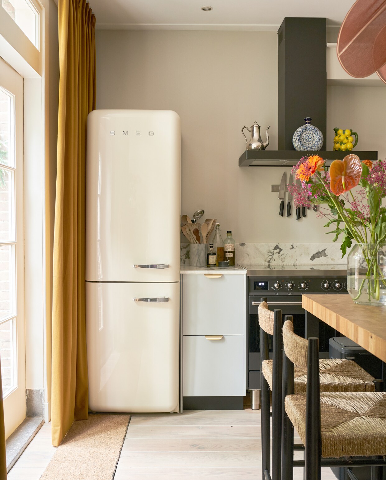 Fornuis en koelkast Smeg, marmeren pot meegenomen uit New York, blauwe karaf uit Mexico, zilveren theepot van Emilie’s grootmoeder, citroenenkaraf &amp;klevering, gordijnen Veneta, barkrukken Zara Home, keuken Veddinge en keukeneiland Ikea, marmeren blad Jan Reek Natuursteen, vaas Bloomon, messing greepjes Luste van Meraki, keukengerei en messenrek onbekend. vtwonen 02-2023 | productie CocoFeatures.com | fotografie Muk van Lil | tekst Caroline Westdijk