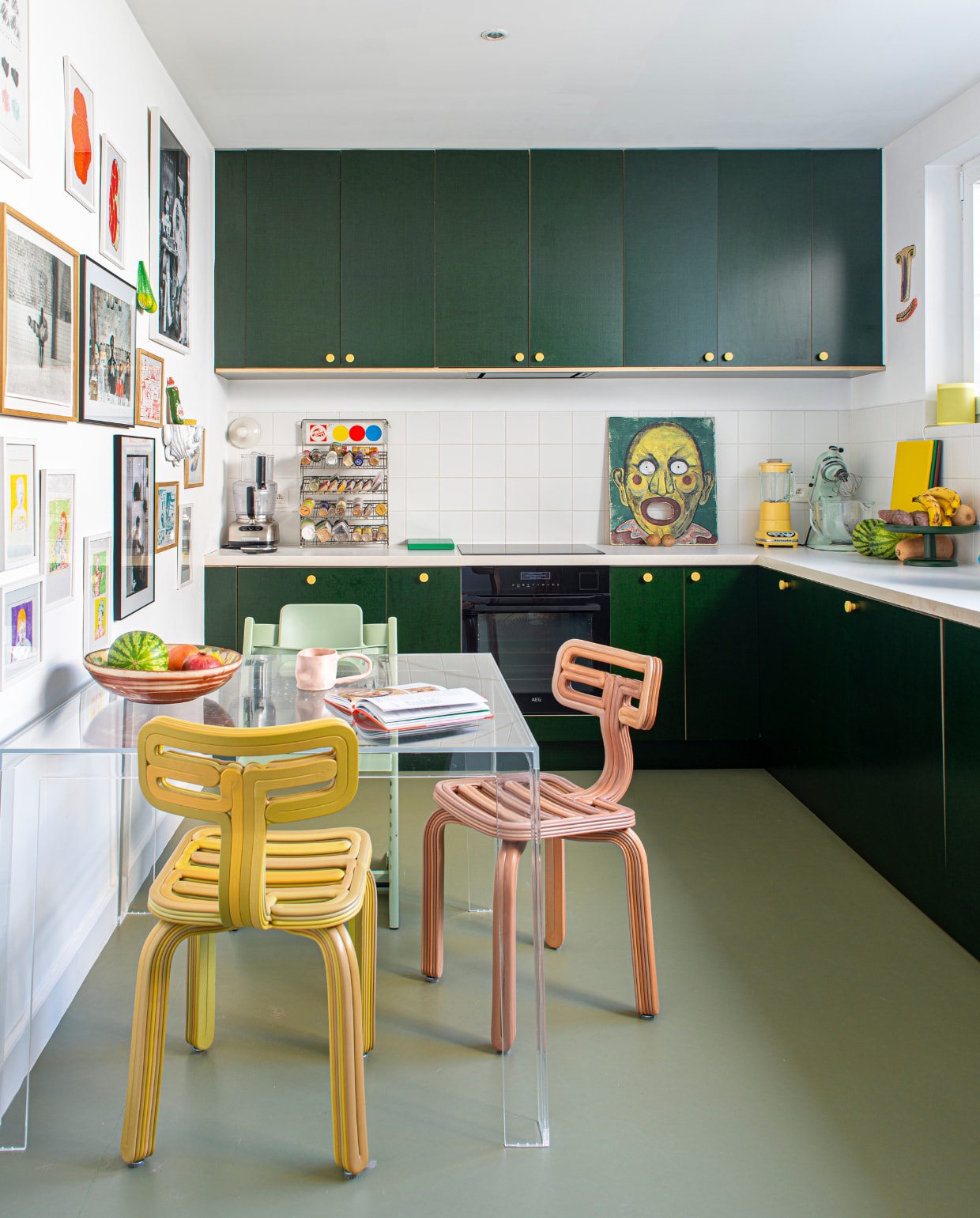 Keukenkastdeuren zelfgemaakt van betonplex met handgrepen van Toniton. Vloer Marmoleum in Rosemary Green van Forbo. Stoelen Chubby van Dirk van der Kooij, tafel Invisible van Tokujin Yoshioka voor Kartell. Blauw object Mommy I made an Ashtray van Rutger de Regt, fruitschaal Vitra, gele speakers Hay x Sonos, naast raam keramisch gezicht Choses Faites Mains, schilderij op aanrecht vintage, spel passe-boules, oud verfdisplayrek van Talens gebruikt voor kruiden. Wandlamp Teti van Artemide. Aan de wand kunstwerken van onder anderen Louise Daneels, Mickey Philips, Hélène Jean-Bon, Joost Stokhof en Mark Bohle. Overig onbekend. vtwonen 02-2023 | styling: Jeske Weel | fotografie: Caroline Coehorst