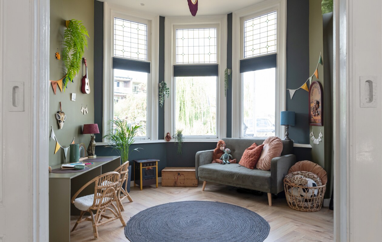 Muurkleur blauw Parcours van Praxis, muurkleur groen Oerwoud van Histor, pvc-vloer lag er al. Bijzettafel, houten fornuis, blauwe tafellamp, plantenpotten en muurbord kringloopwinkel. Kist en bank Marktplaats. Bruine aap en mand Ikea, blauwe knuffel en korte slinger Hema, kussens v.l.n.r. Blokker en MrsBloom. Lange slinger Savanna van Nobodinoz, gitaar New Classic Toys, kunsthangplant Asparagus van Easyplants. Tafellamp Xenos, stoelen Dora via Casa, tegels StoryTiles, giraffe Melissa &amp; Doug, abacus Little Dutch, blik Zeeman, planten Intratuin, vloerkleed Kwantum. Bureau onbekend. vtwonen 02-2023 | Styling Sharon Roest | Fotografie Caroline Martinot | Tekst Mirjam Rosema-Verhulst