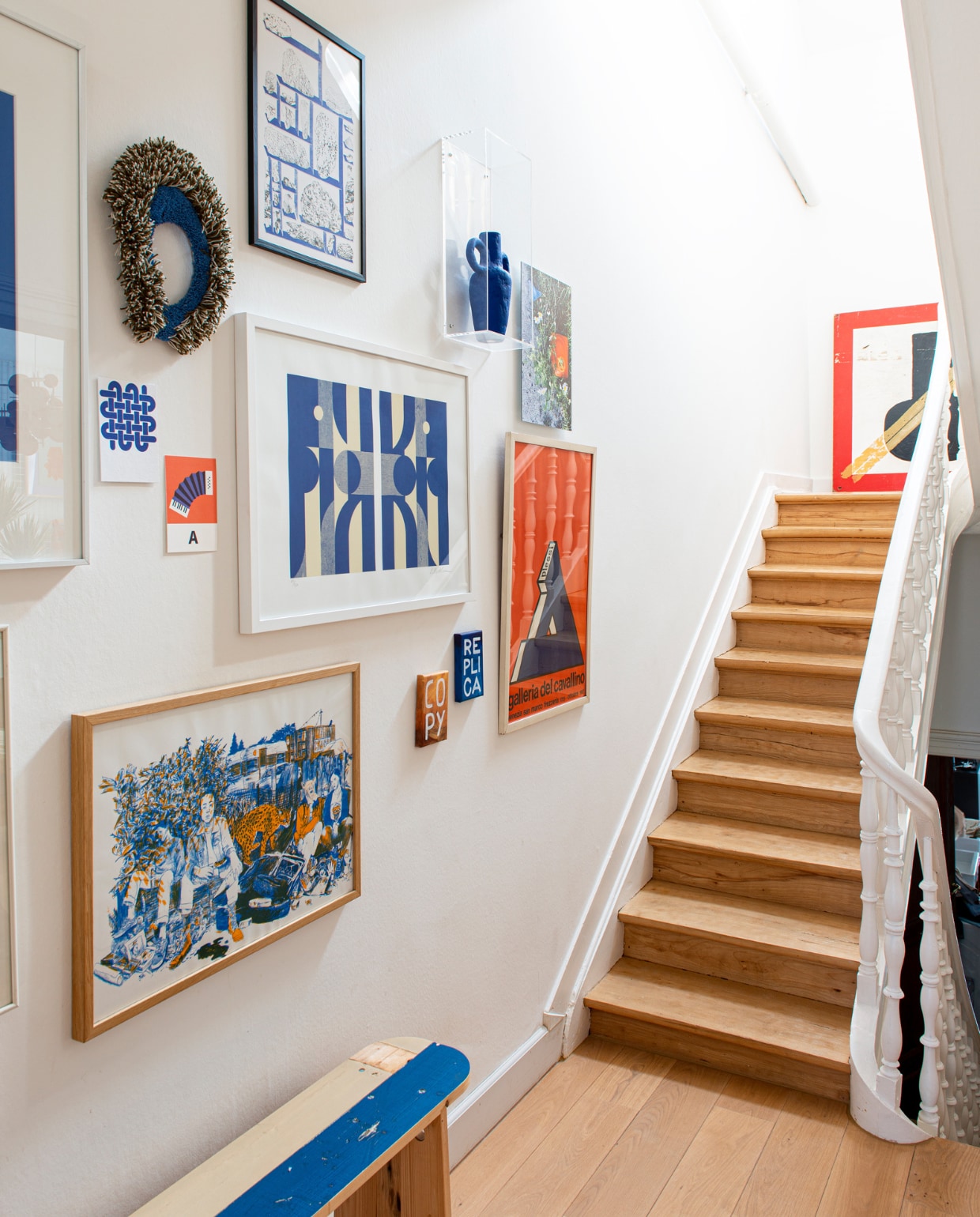 Bank Piet Hein Eek, daarboven risoprint Ward Zwart, ernaast twee kleine schilderijen van Jan Leenders, in het midden blauw-witte zeefdruk van Louis Reith, links ernaast prints Edouard Schneider, erboven rond getuft object Kihnu, helemaal boven risoprint Mélanie Corre, rechts daarvan vaas papierklei Jacqueline de la Fuente, daarnaast foto van Eline, eronder typografisch affiche tentoonstelling Plessi, bovenaan de trap een houten signaalbord voor de scheepvaart. vtwonen 02-2023 | styling: Jeske Weel | fotografie: Caroline Coehorst