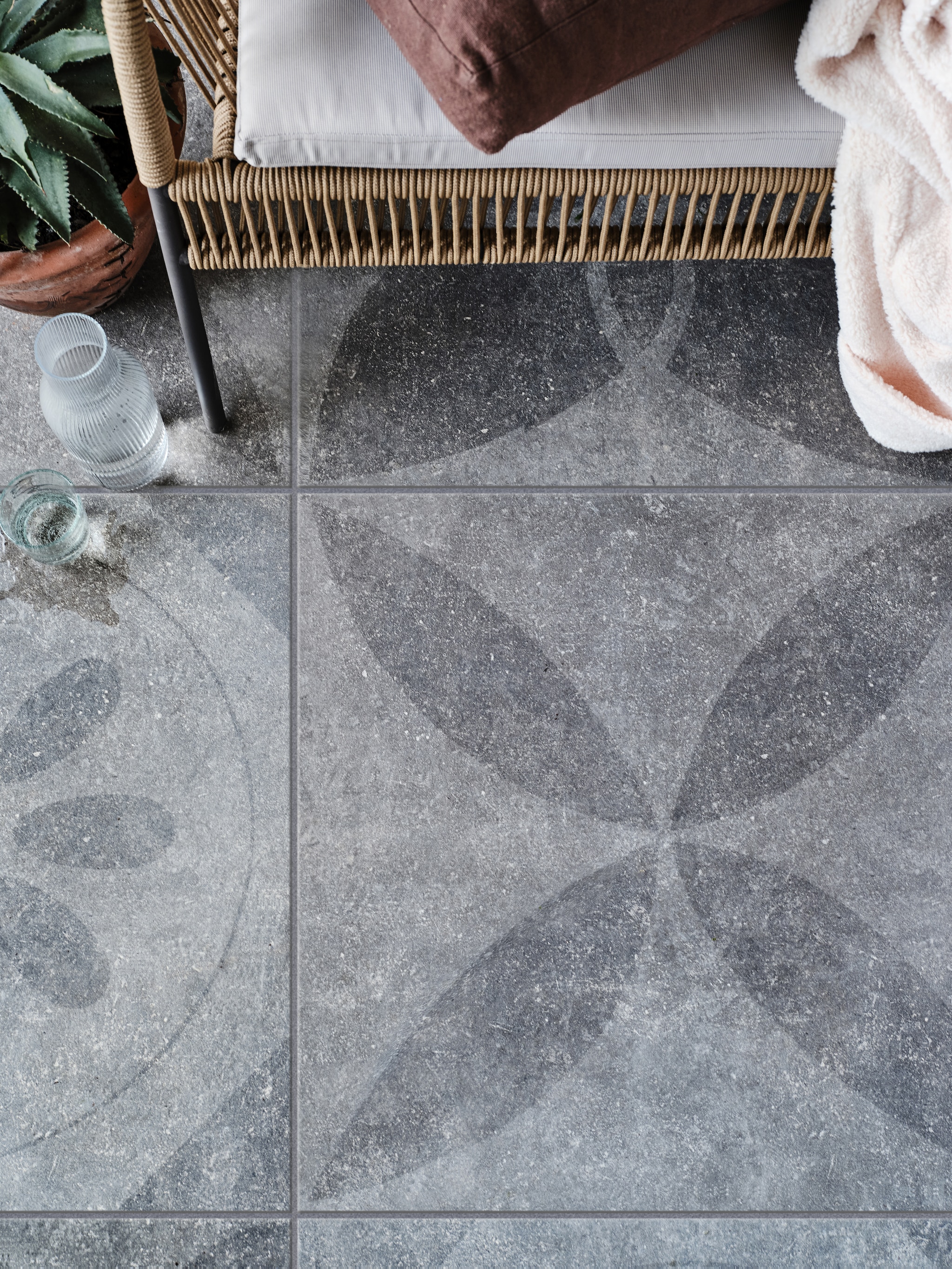 Tuintegel Solostone mix van verschillende dessins: Hormigon Butterfly Antracite, 70x70 cm, per m² € 79,- + Hormigon Floret Antracite, 70x70 cm, per m² € 79,-, Hormigon Flower Antracite, 70x70 cm, per m² € 79,- (ook in 90x90 cm, per m² € 89,50), Hormigon Citrus Antracite, 70x70 cm, per m² € 79,-. M.m.v. vtwonen.nl (tweezits tuinbank Samantha van Kave Home, glas en karaf van Serax), householdhardware.nl (tadelakt bloempot, kussen), jolandelijk.nl (opbergkist, doosjes, kapstok, houten spijlenstoel). vtwonen 03-2023 | styling Marlies Does | fotografie Bart Brussee