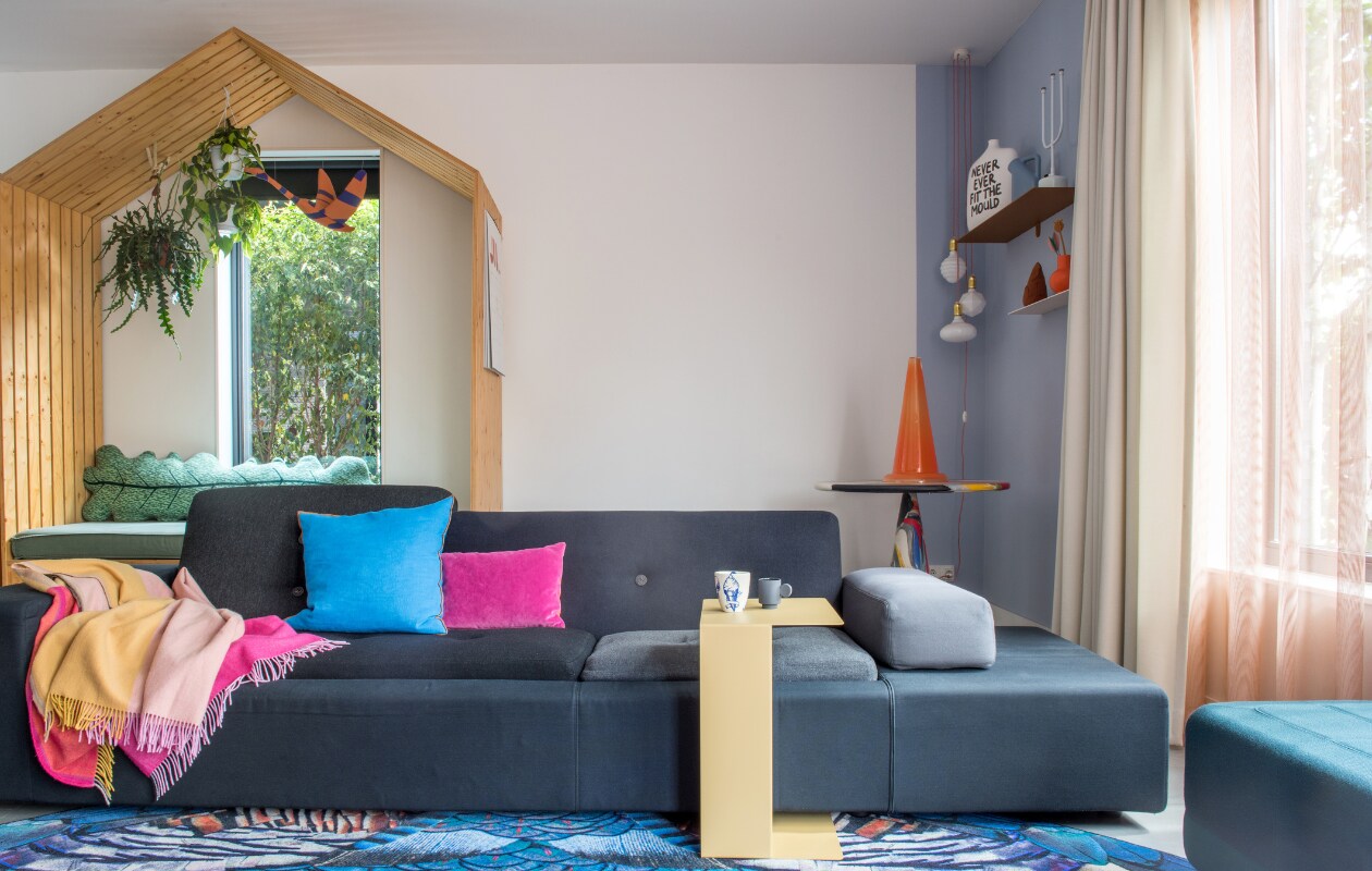 Houten huis eigen ontwerp by Maykel, bladkussen Donna Wilson, zwaan Jantien Baas. Zetel Polder Sofa en plaid Hella Jongerius voor Vitra, vloerkleed Moooi Carpets, blauw kussen Hay, paars kussen Nijhof, bijzettafel Gelderland Ori, kopje links Social Label, rechts Maarten Baas, poef Normann Copenhagen. Tafel in hoek Dirk Vander Kooij, kunstwerk Save me Glass traffic cone van Bas Kosters, wandplanken Muller van Severen, vaas Irenic Mould van Birgit Verwer via Cor Unum, karaf Muuto, kandelaar Chris Kabel voor Droog Design, duif Christien Meindertsma, oranje vaas Raawii, lampen Zangra. Overgordijnen op maat gemaakt door stoffeerder, vitrage van Kvadrat-stof, kleur op muur onbekend. vtwonen 02-2022 | productie Anke Leunissen | styling Kim de Groot | fotografie Anke Leunissen