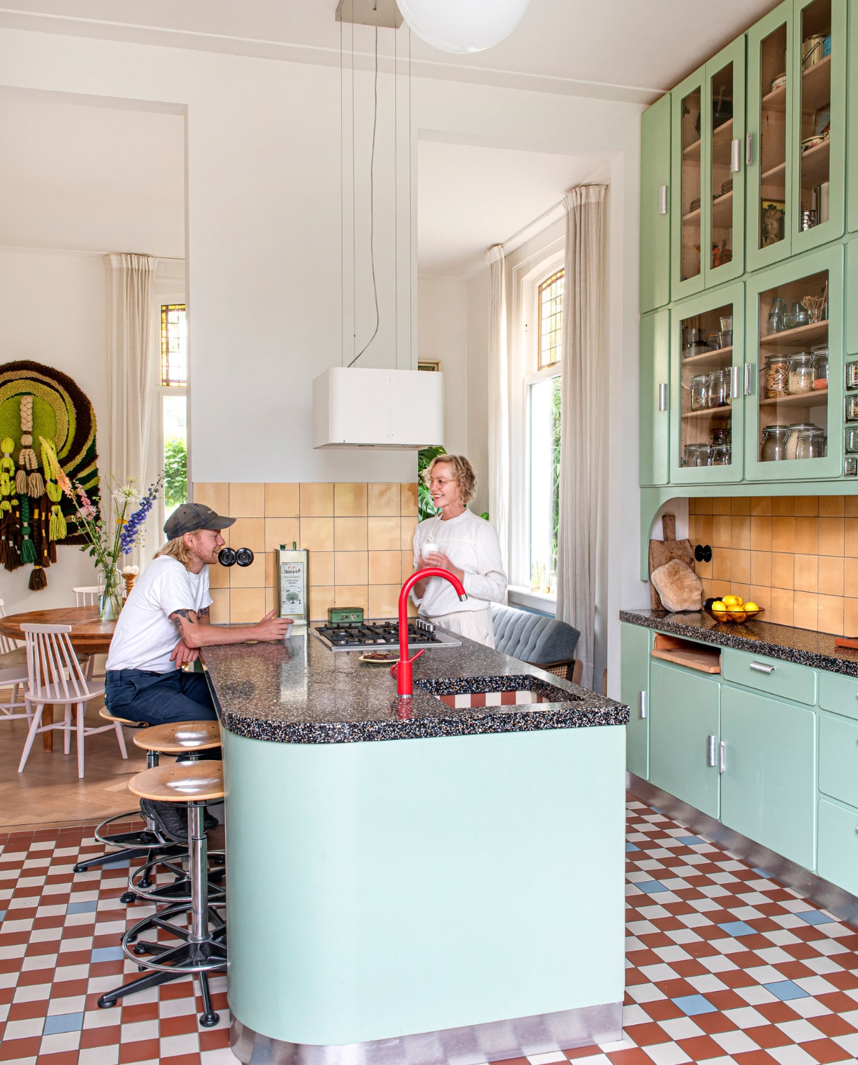 Keuken ontworpen door Studio Daan Hietbrink, verfkleur fronten Room Green 81 van Farrow &amp; Ball. Keukenblad Marcolina Terrazzo en Beton. Rode kraan sanitair.nl. Wandtegels en barkrukken Marktplaats. Vloertegels Winckelmans. Fruitschaal en broodplank uit Frankrijk. Overig onbekend. vtwonen 03-2023 | Styling en fotografie Caroline Coehorst i.s.m. Studio Daan Hietbrink