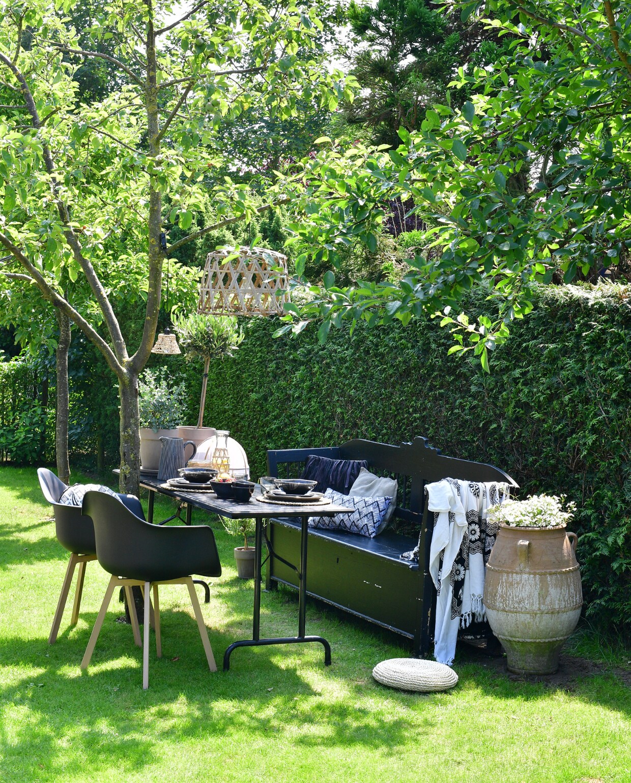In de tweede tuinkamer is een zit- en eetplek ingericht onder de appelboom. Klepbank overgenomen van een kennis en zwart geverfd. Zwart geverfde picknicktafel gemaakt van een onderstel van de rommelmarkt en een zelfgetimmerd blad. Stoelen, lampen, plaid, kussens en kan Loods 5. Grote pot tuincentrum. Karaf, placemats, glas, bestek en  muggennet Xenos. Servies Broste Copenhagen. Potten met olijf- en eucalyptusboompje TP Flowers &amp; More. vtwonen tuinspecial tuin 7 2023 | fotografie sietske de vries