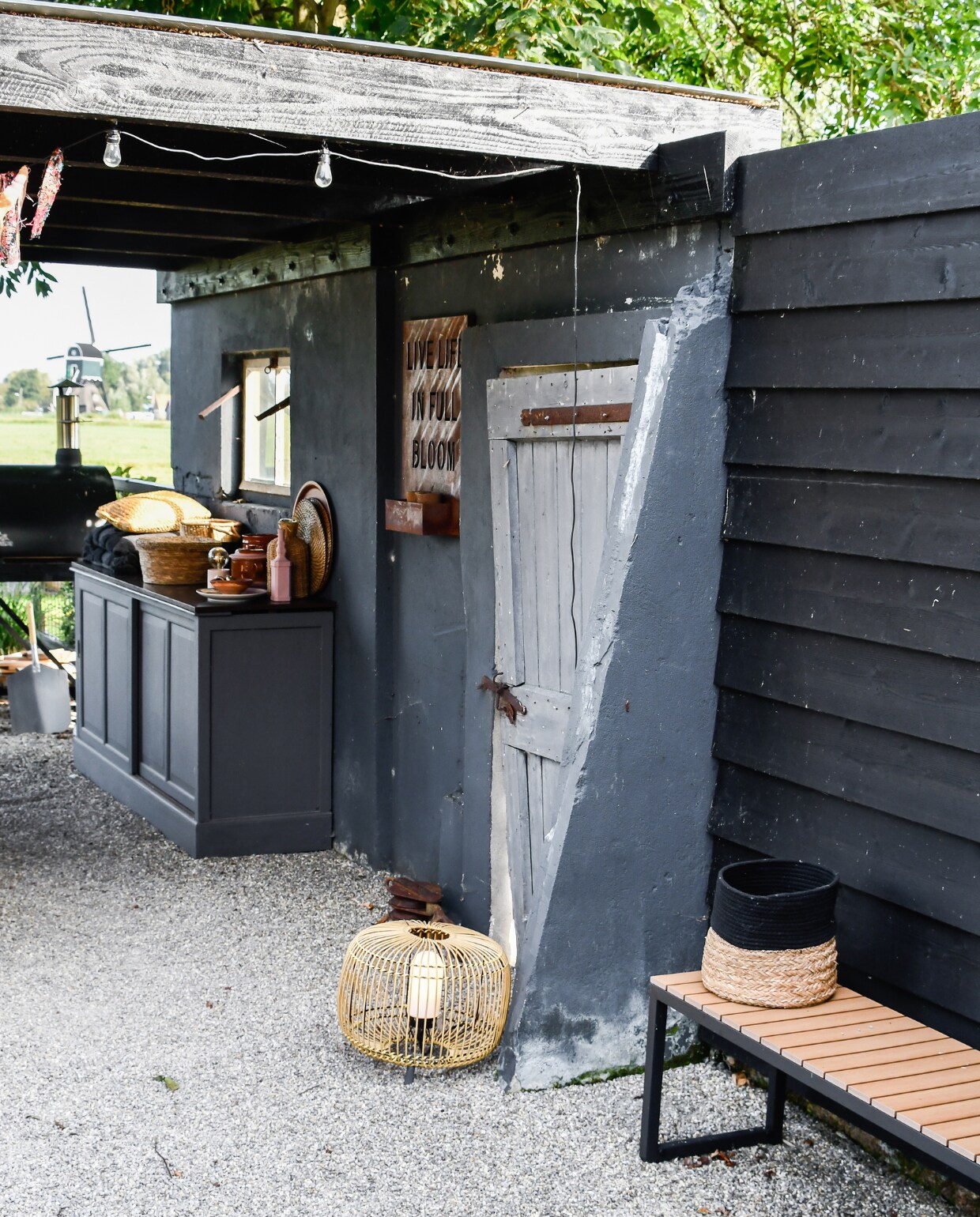 De overkapping van het terras rust op de muur van de oude geitenstal met de oorspronkelijke deur. Het kastje was ooit in gebruik als buffetkast, de bovenkant ging naar de stort, de onderkant heeft Saskia met grijze muurverf geschilderd. Lamp op de grond Kwantum, bank en lampje Jysk, verroeste tekst aan de muur Intratuin. Op de kast fles geverfd met roze muurverf, ronde mand met deksel Kwantum, alle potten kringloop, kussens Loods 5. Het terras van grind lag er al. Het voordeel van grind is dat het goedkoop is en voor een goede afwatering zorgt. Ook krijgt onkruid minder kans. vtwonen special tuin 07 | styling Marieke de Geus | fotografie Barbara Groen