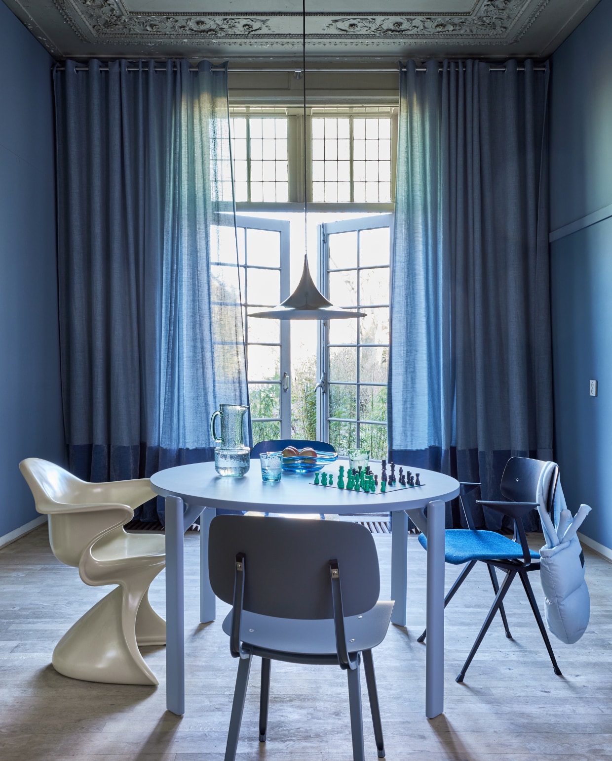 Blauwe gordijnstof Tick-Tock Crushed Reboot van Chivasso, linnen en polyester, kamerhoog 300 cm, per meter € 147,50, lichtblauwe fluwelen stof Sensual Velvet van Chivasso, polyester en katoen,140 cm breed, per meter € 105,- (jab.de). Rvs gordijnroede inclusief steunen en eindknoppen, per meter vanaf € 115,- (xxldesignroeden.nl). Krijtverf Classico in Blue Stone, per liter € 55,75 (pure-original.com). Houten tafel Rey, 128,5x74,5 cm (Øxh) € 2049,-, blauwe eetkamerstoelen Revolt € 385,-, Result € 509,- en Rey € 549,-, kartonnen schaakspel van Hay € 55,-  (hay.nl). Hanglamp € 490,-(goeds.nl). Roomgele plastic stoel Sedia Sabbia € 895,- (hkliving.nl). Tas € 99,95 (madsnorgaard.com). Gekleurde drinkglazen, set van 6 € 119,-, glazen schaal Eye € 125,-, glazen waterkan € 62,50 (polspotten.com). vtwonen 05-2023 | styling Marianne Luning | fotografie Alexander van Berge